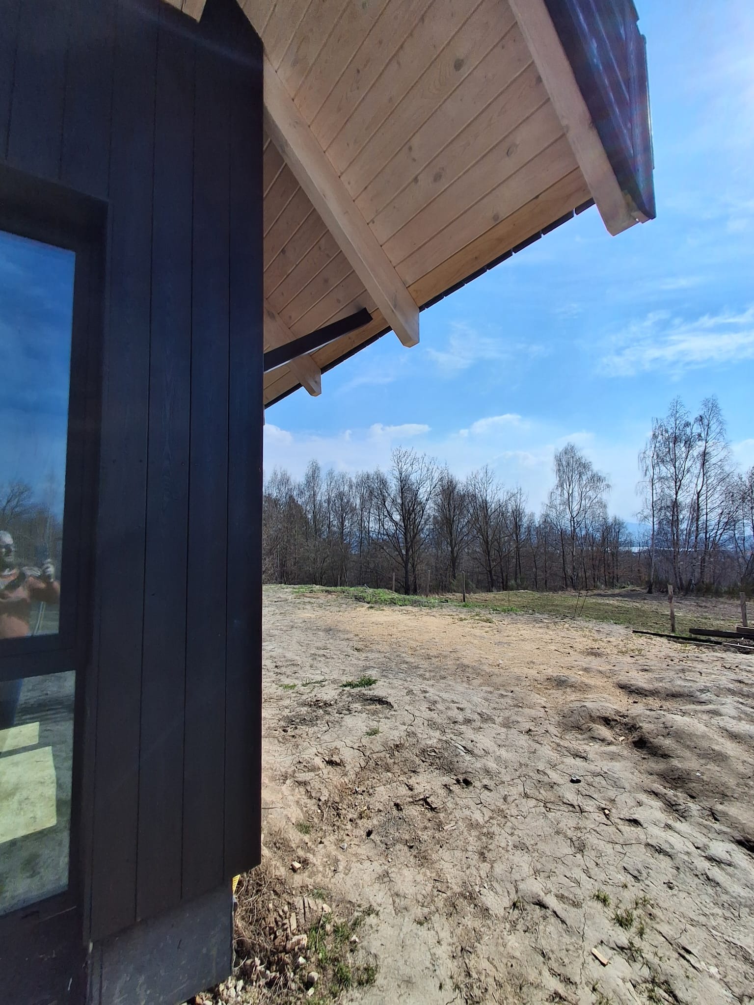 Ciemna elewacja domu modułowego z drewnianym zadaszeniem, widok na krajobraz z drzewami i błękitnym niebem w tle. Surowy teren wokół budynku.