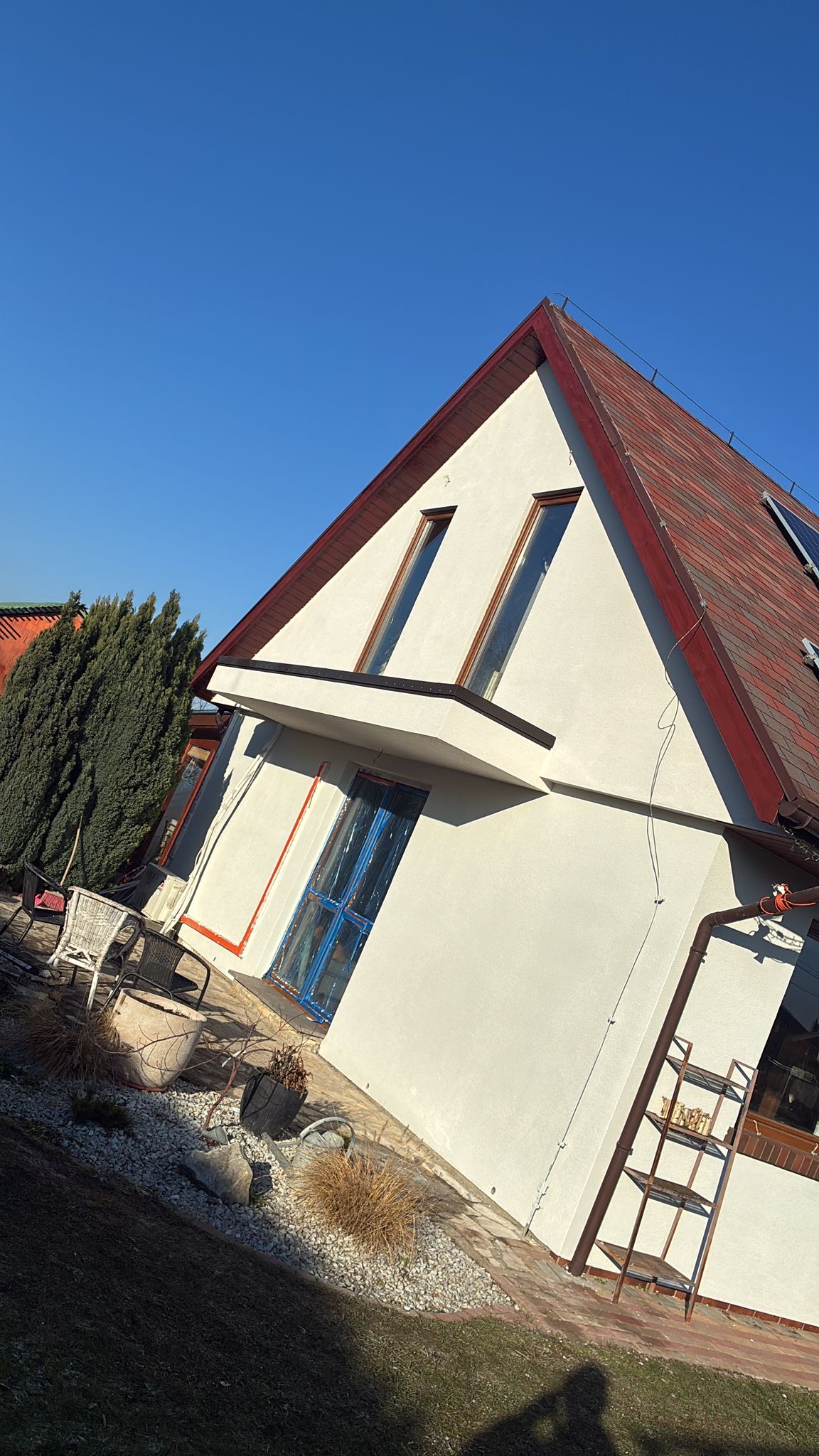 Biała elewacja domu z czerwonym dachem i oknami w brązowych ramach. Okno na parterze zabezpieczone folią. Widoczne elementy ogrodu i drabina.