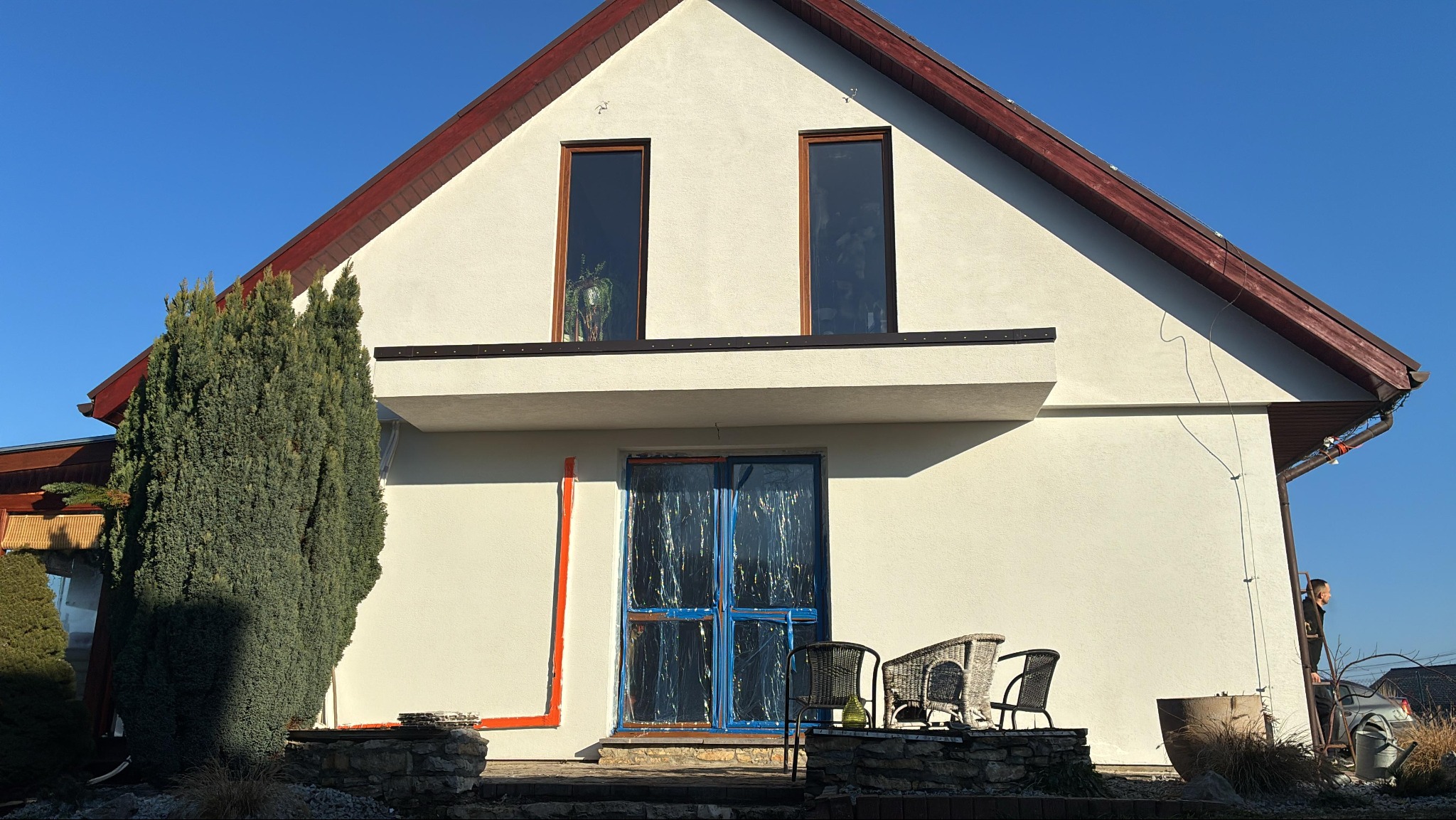 Biała elewacja domu z czerwonym dachem i wysokimi oknami. Drzwi zabezpieczone folią malarską, widoczne ślady po malowaniu. Krzesła ogrodowe na tarasie.