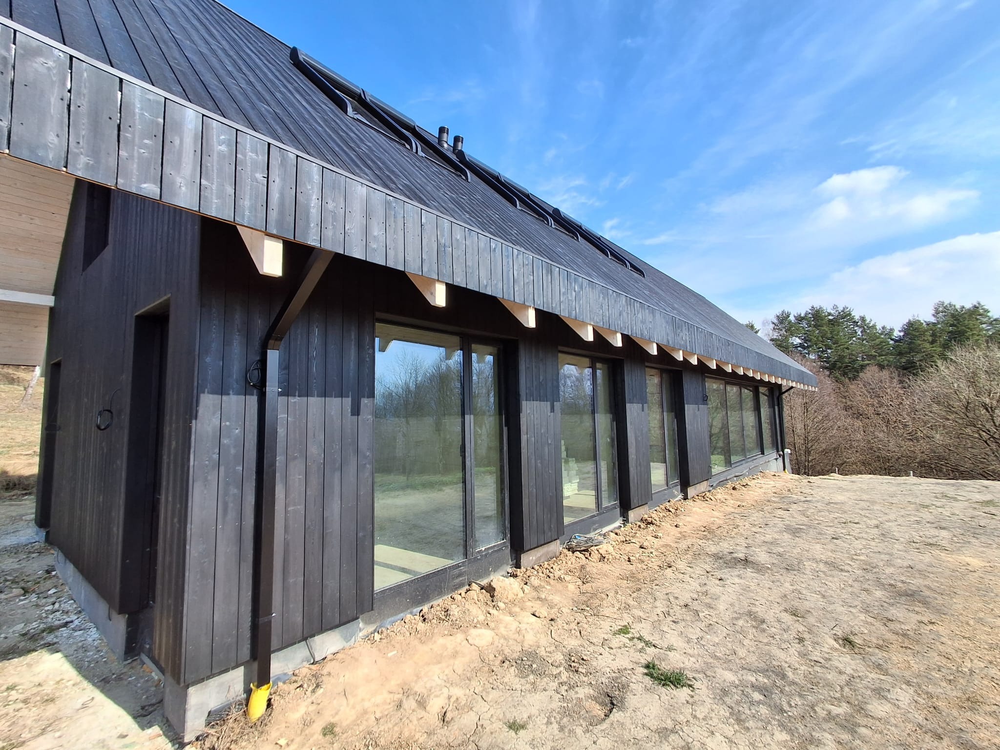 Nowoczesny dom modułowy z czarną drewnianą elewacją i dużymi oknami, widok z zewnątrz w słoneczny dzień. Charakterystyczne drewniane wsporniki pod okapem.