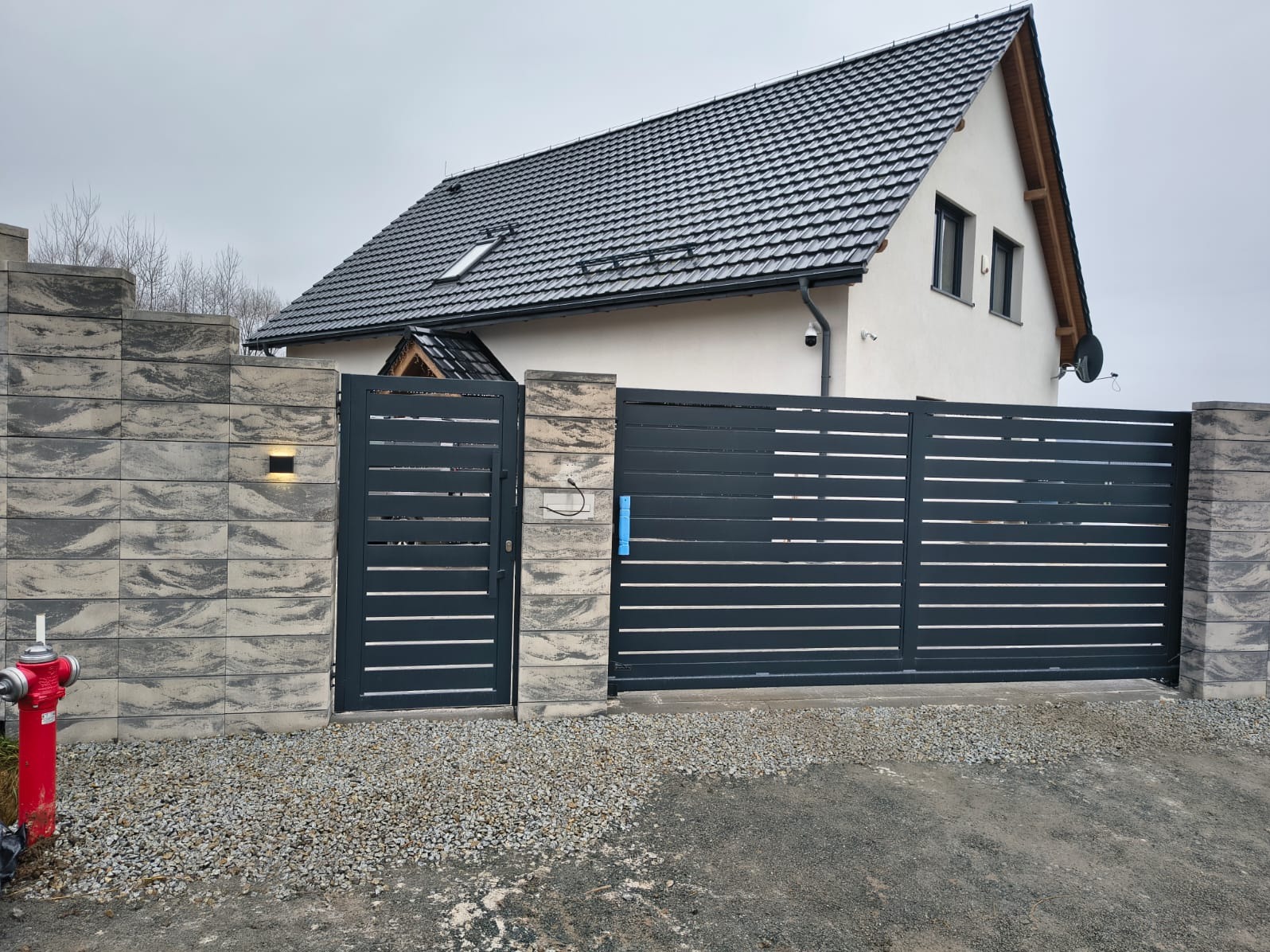 Nowoczesna, przesuwna brama w kolorze antracytowym z poziomymi panelami, wkomponowana w mur z szarej kostki. Dom z ciemnym dachem w tle.
