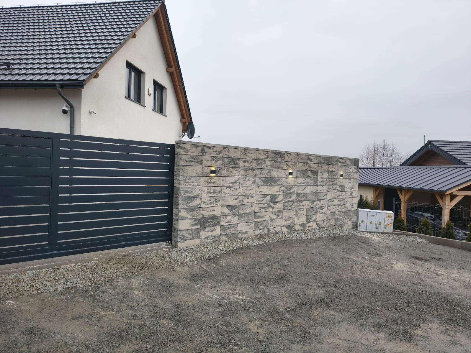 Nowoczesna, pozioma brama wjazdowa z szarymi panelami, przylegająca do muru z dekoracyjnej szarej cegły z oświetleniem. Dom w tle, pochmurne niebo.