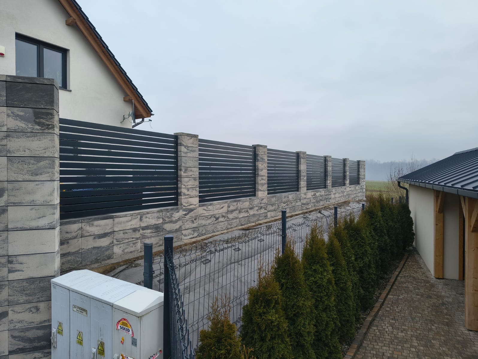 Nowoczesne ogrodzenie panelowe z poziomymi, czarnymi przęsłami i kamiennymi słupkami, widok z perspektywy posesji, zieleń wzdłuż ogrodzenia, pochmurne niebo.