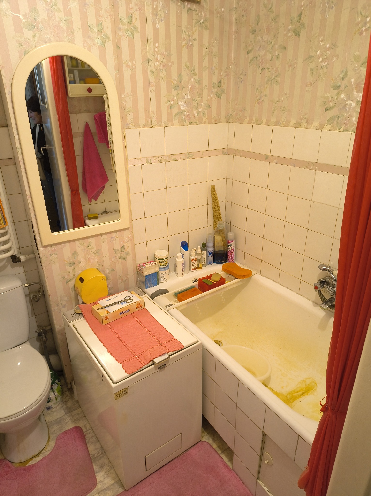 Zaniedbana łazienka z wanną, pralką i toaletą. Na pralce leżą nożyczki i pudełko. Widoczne płytki i tapeta w kwiaty. W lustrze odbija się fragment pomieszczenia.
