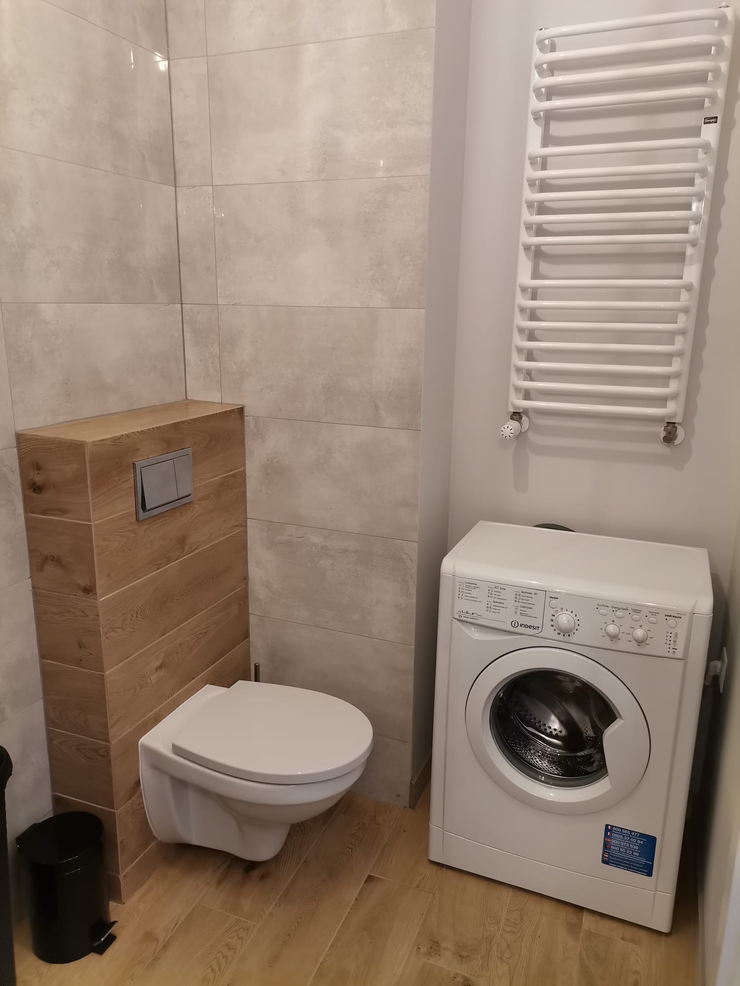 Jasna łazienka po remoncie: podwieszana toaleta z drewnianą obudową, grzejnik drabinkowy i pralka Indesit. Płytki w odcieniach szarości i beżu.