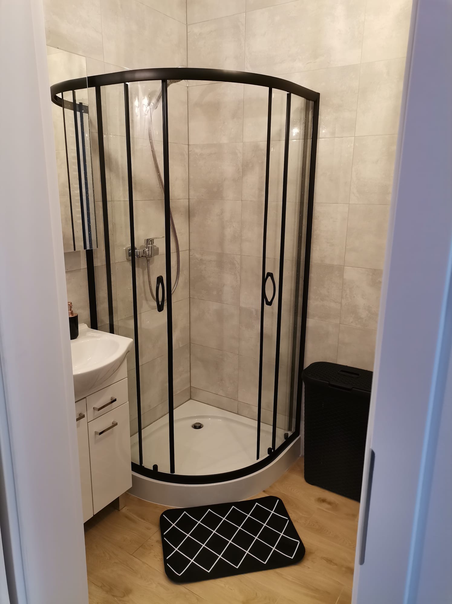 Nowoczesna kabina prysznicowa z czarnymi profilami, biała umywalka z szafką, szara płytka na ścianie i drewnopodobna podłoga. Czarny kosz na pranie i dywanik.