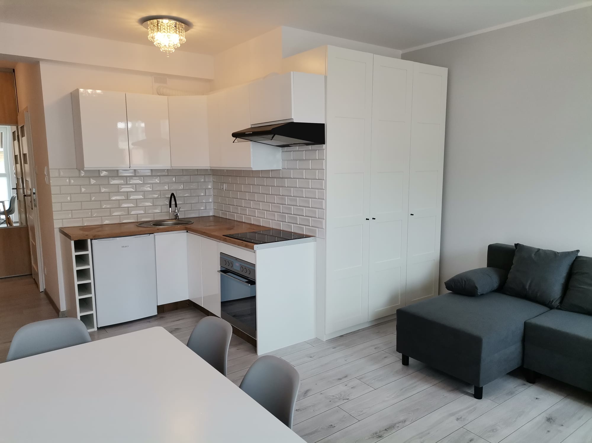 Jasna kuchnia z białymi meblami, szarą cegłą na ścianie i drewnianym blatem. Widoczny stół z krzesłami i szara sofa. Całość w nowoczesnym stylu.