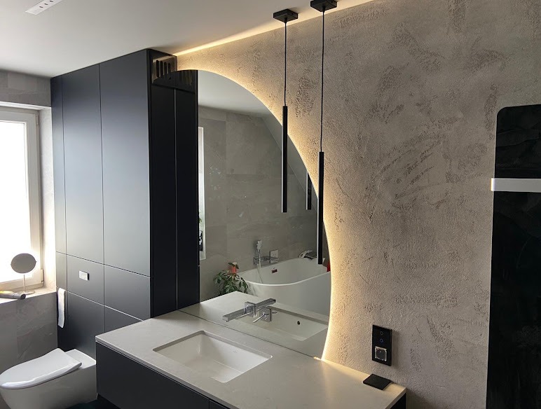 Nowoczesna łazienka z podświetlanym lustrem w kształcie półksiężyca, czarnymi szafkami i wiszącymi lampami. Dekoracyjna ściana z efektem betonu.