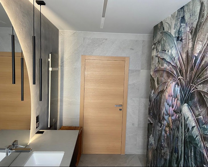 Nowoczesna łazienka z drewnianymi akcentami: drzwi, szafka pod umywalką i lustro z podświetleniem LED. Na ścianie tapeta z motywem roślinnym i szare płytki.