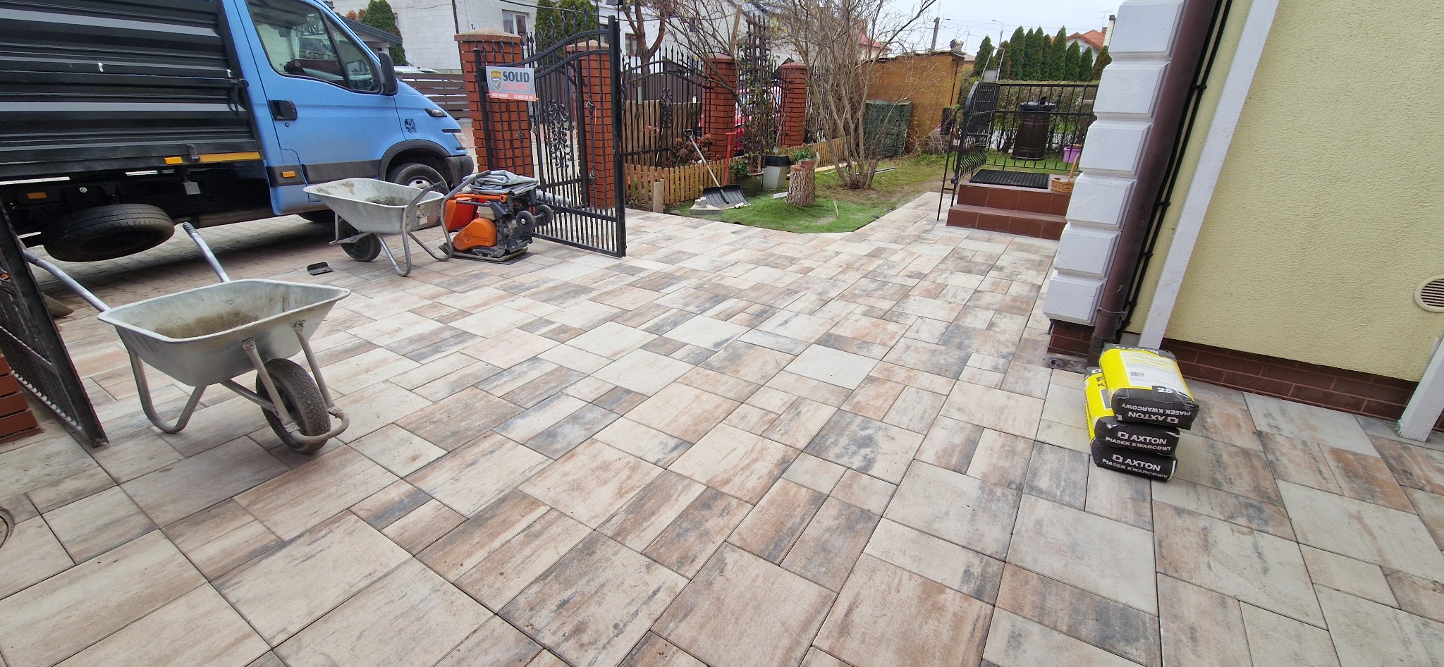 Ułożona kostka brukowa w odcieniach beżu i szarości, z widocznymi narzędziami: wozidła, zagęszczarka. W tle brama wjazdowa i zaparkowany samochód dostawczy.