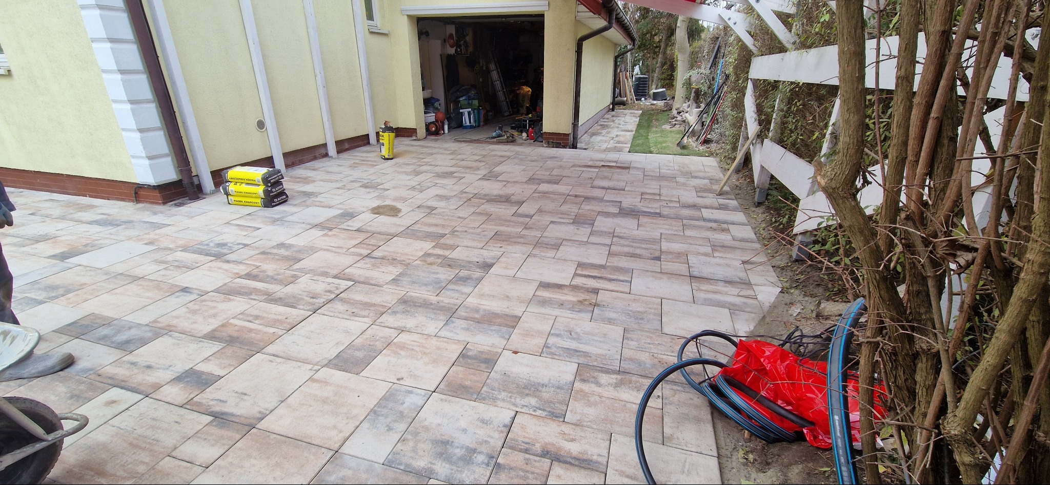 Ułożona kostka brukowa w odcieniach beżu i szarości prowadzi od garażu do ogrodu. Widoczne worki z piaskiem i narzędzia. Całość w neutralnej tonacji.