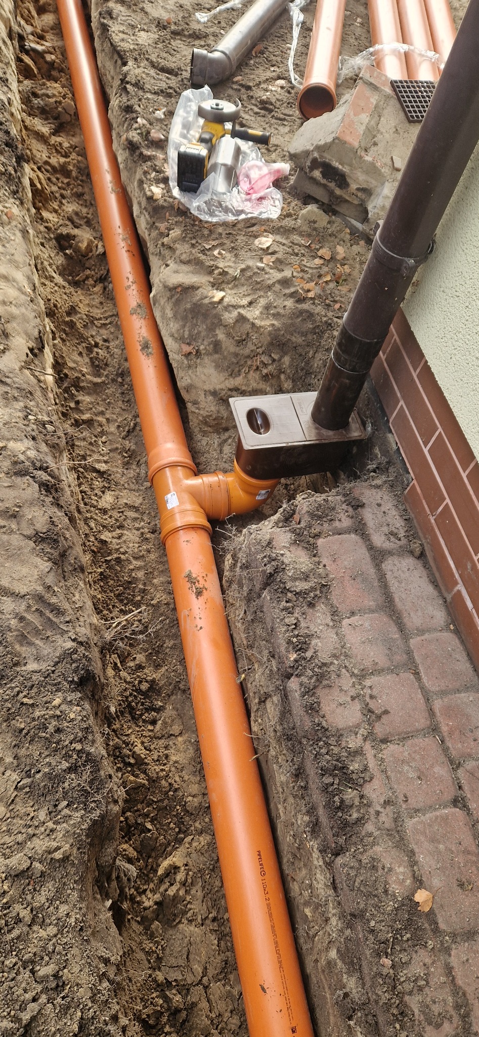 Pomarańczowa rura drenażowa ułożona w wykopie ziemnym, połączona z odpływem rynnowym. Widoczne narzędzia i fragment kostki brukowej. Ujęcie z góry.
