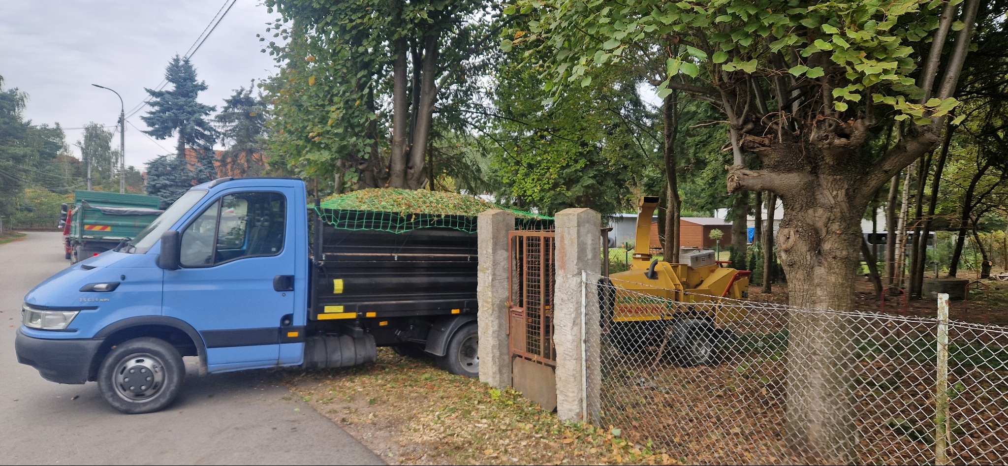 Niebieska ciężarówka z przyczepą wypełnioną zielenią, obok żółta maszyna do rozdrabniania gałęzi za ogrodzeniem. W tle drzewa i budynki.