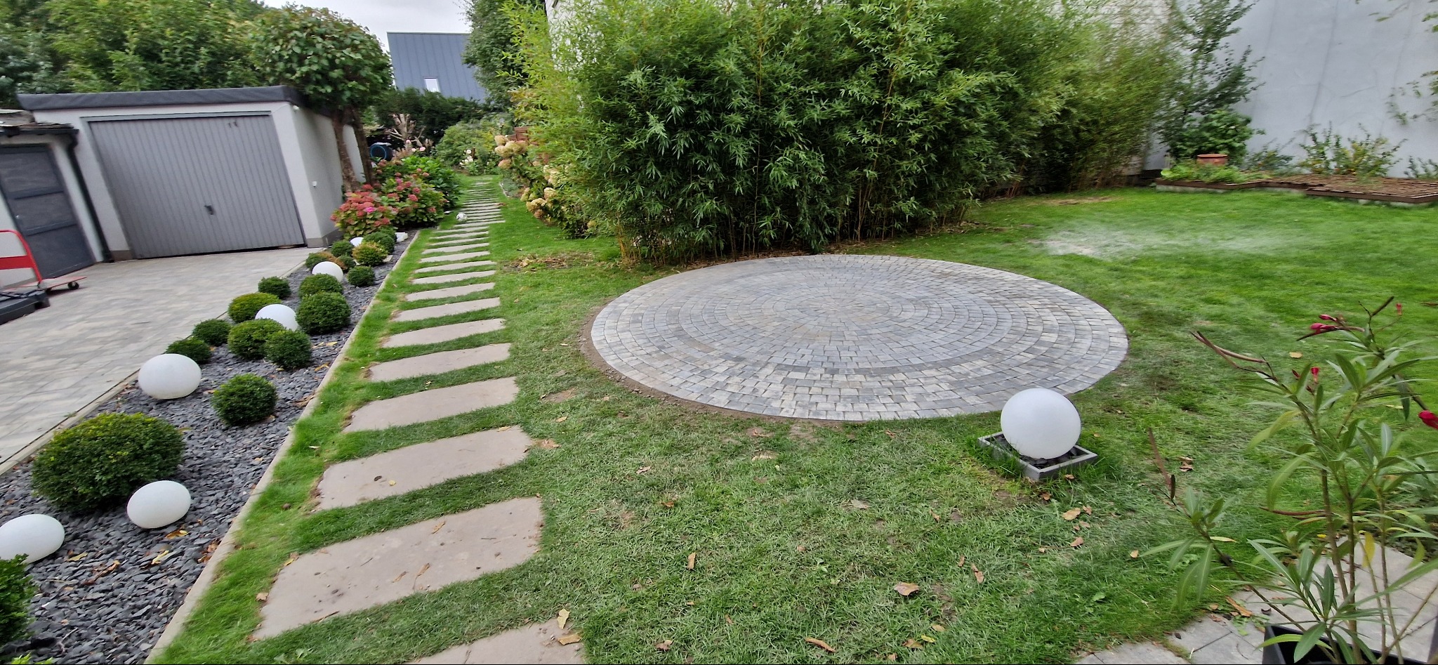 Ogród z okrągłym placem z kostki brukowej, ścieżką z płyt i dekoracyjnymi kulami. Widok na zadbany trawnik, krzewy i garaż w tle. Całość tworzy harmonijną przestrzeń.