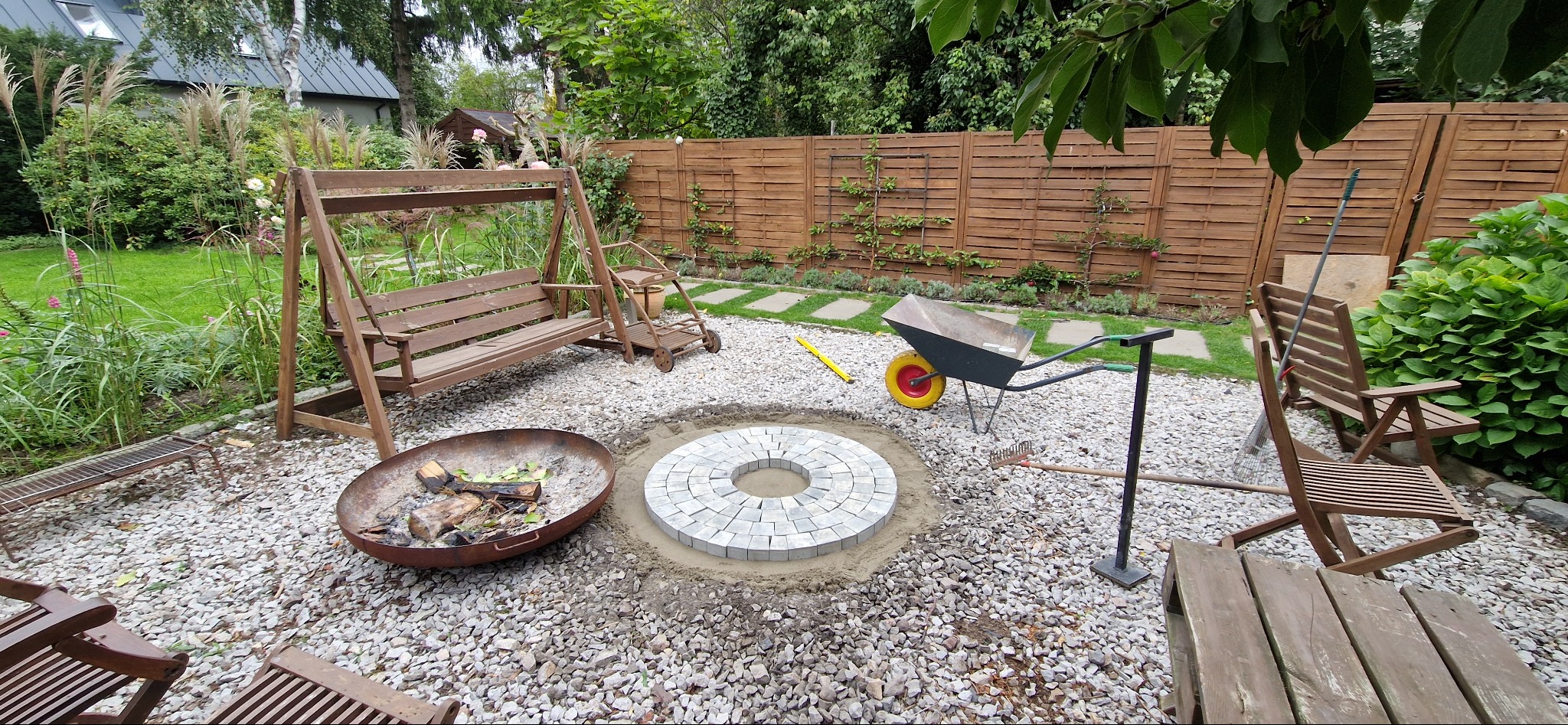 Ogród z paleniskiem z kostki brukowej w trakcie budowy, otoczony żwirem, z drewnianą huśtawką i meblami ogrodowymi. W tle drewniany płot i zieleń.
