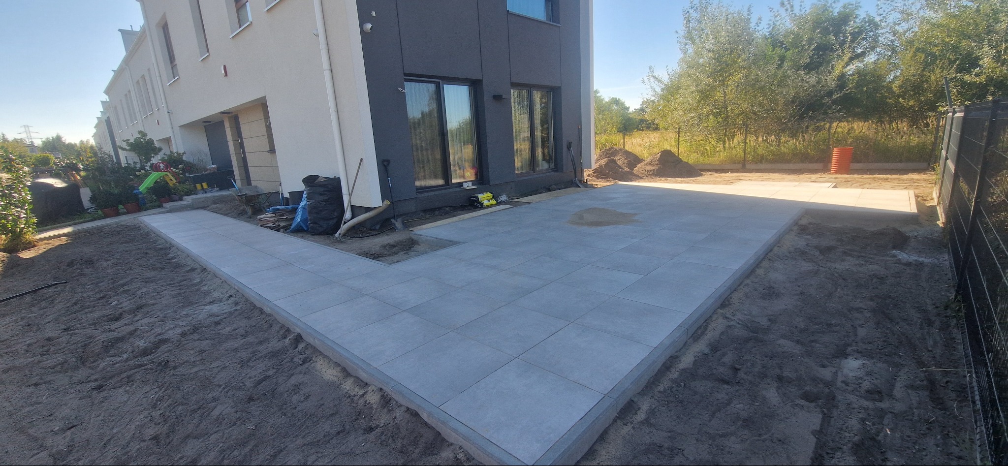 Ułożona, szara kostka brukowa na tarasie przydomowym, widok z perspektywy ziemi. Częściowo wykończony teren, widoczne narzędzia i worki budowlane przy budynku.
