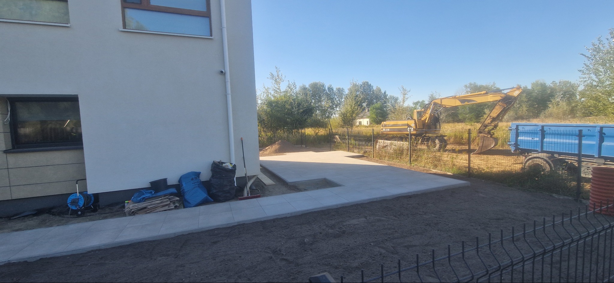 Fragment ułożonej, szarej kostki brukowej przy budynku. W tle widoczny żółty koparka i niebieska wywrotka. Prace ziemne w toku, teren przygotowany pod dalsze prace.