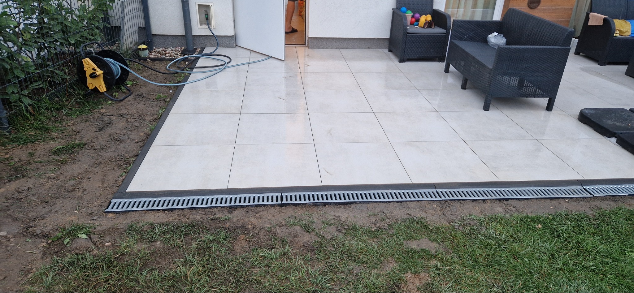 Jasne płytki tarasowe z odpływem liniowym, otoczone ziemią i trawą. W tle meble ogrodowe i częściowo otwarte drzwi.