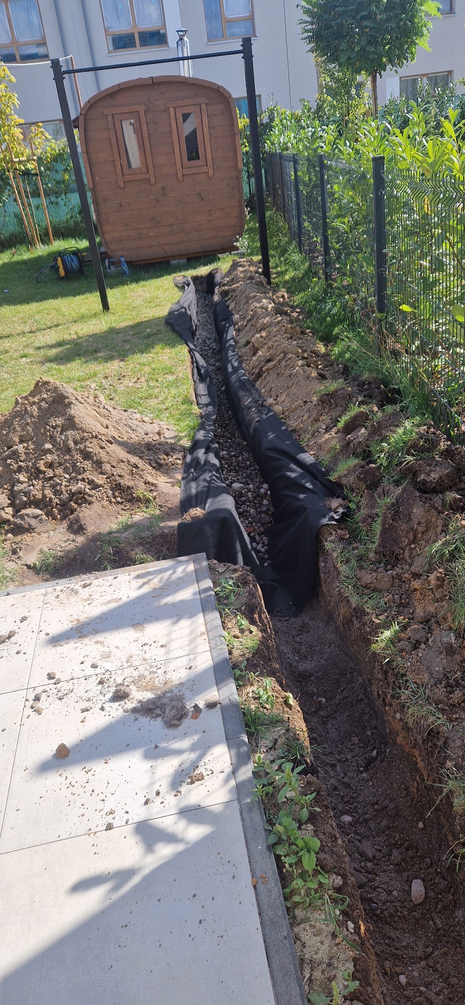 Wykopany rów z czarną geowłókniną i kamieniami, prawdopodobnie drenaż, obok kostka brukowa i trawnik. W tle domek dla dzieci i ogrodzenie.