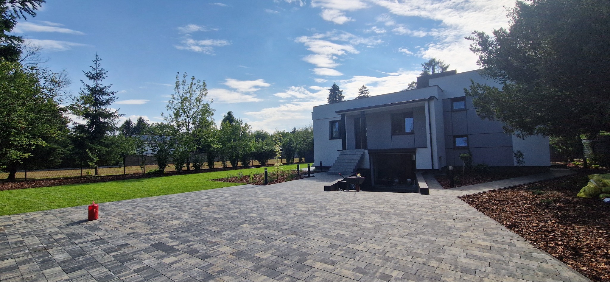 Nowoczesny dom z szarą kostką brukową, zielonym trawnikiem i ozdobnymi nasadzeniami. Widok na podjazd, budynek i fragment ogrodu w słoneczny dzień.