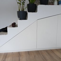 Stolarnia Gwiaździsta Grzegorz Kazanowski - Białe szafki pod schodami z roślinami w doniczkach na stopniach, drewniana podłoga. Funkcjonalne wykorzystanie przestrzeni pod schodami.