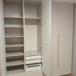 Stolarnia Gwiaździsta Grzegorz Kazanowski - Jasna szafa wnękowa z otwartymi drzwiami, ukazująca półki, drążek na ubrania i szuflady. Dolna część szafy zdobiona pionowymi żłobieniami. Widoczna podłoga z paneli.