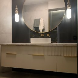 Stolarnia Gwiaździsta Grzegorz Kazanowski - Nowoczesna łazienka z owalnym lustrem w złotej ramie, białą umywalką nablatową i szafką z frezowanymi frontami oraz złotymi uchwytami. Minimalistyczne oświetlenie.