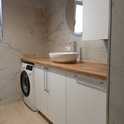Stolarnia Gwiaździsta Grzegorz Kazanowski - Nowoczesna łazienka z drewnianym blatem, białą umywalką nablatową i szafkami. Obok pralka, a na ścianie lustro w okrągłej ramie i marmurowe płytki.
