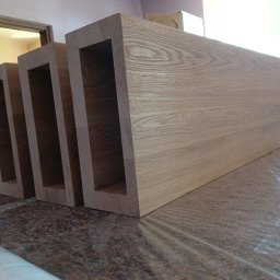 Stolarnia Gwiaździsta Grzegorz Kazanowski - Drewniane, surowe elementy konstrukcyjne z wyciętymi otworami, ułożone jeden za drugim na blacie, w tle widoczne drzwi i grzejnik.