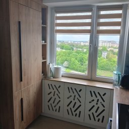 Stolarnia Gwiaździsta Grzegorz Kazanowski - Drewniana szafa z czarnymi uchwytami obok okna z roletami dzień-noc i zabudową grzejnika z ażurowym wzorem. Widok na zieleń miejską za oknem.