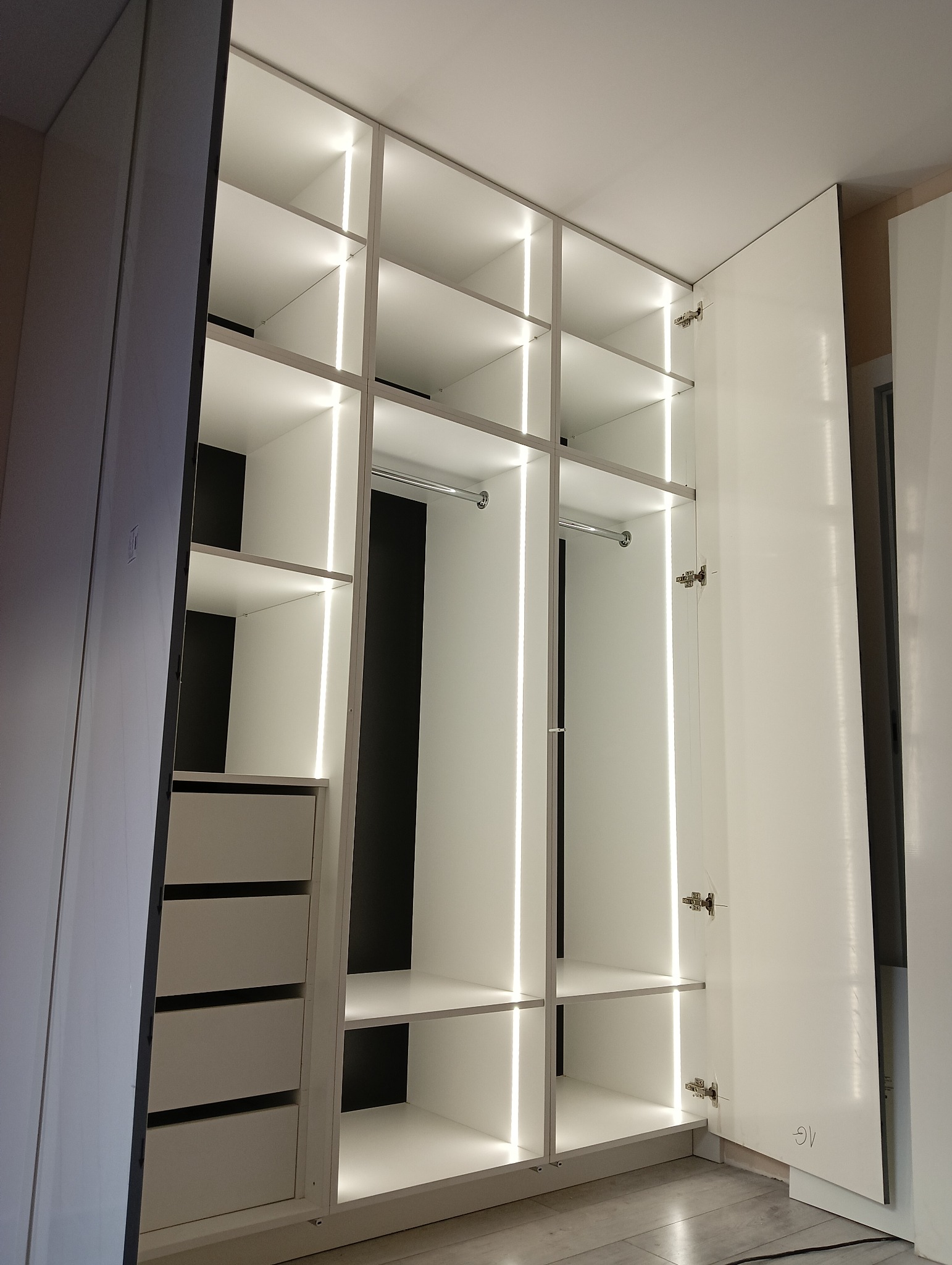 Biała szafa wnękowa z oświetleniem LED, widok z dołu, otwarte drzwi ukazują półki i szuflady. Minimalistyczny design, nowoczesne wykończenie.