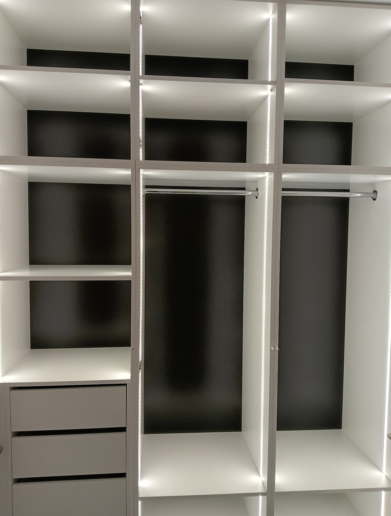 Biała szafa wnękowa z szufladami i oświetleniem LED, widok z bliska na puste półki i drążki na ubrania. Minimalistyczny design z czarnym tłem wewnątrz.