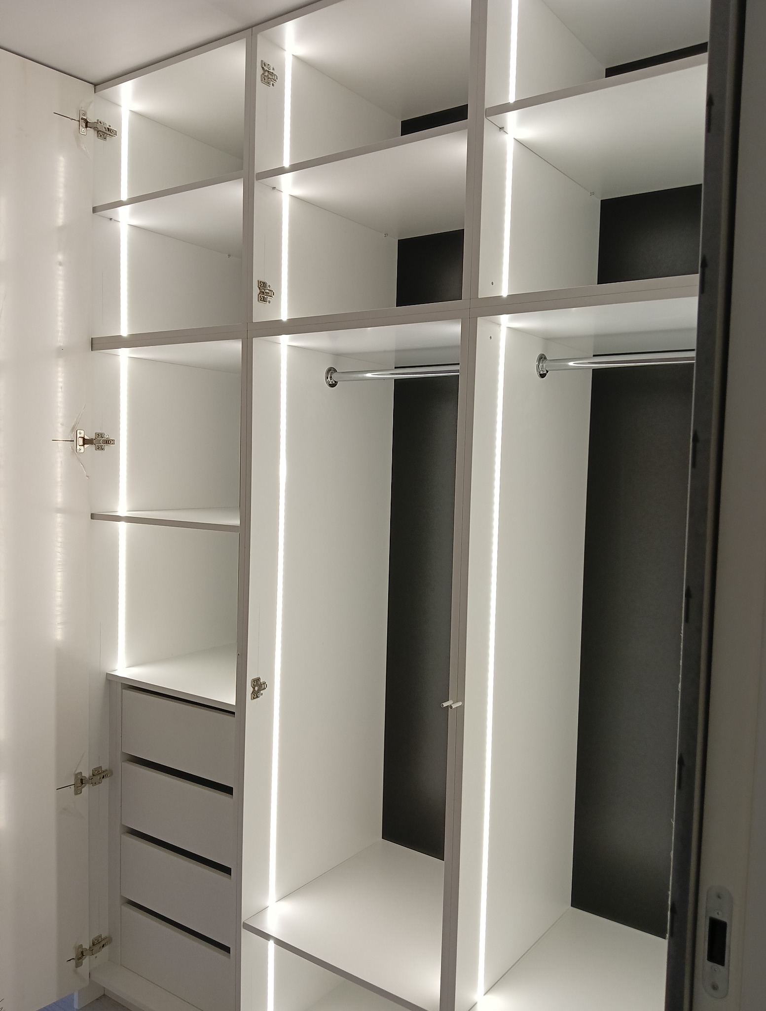 Wnętrze szafy wnękowej z oświetleniem LED, widoczne półki, szuflady i drążek na ubrania. Minimalistyczny design, białe fronty, szare szuflady, czarne tło w głębi szafy.