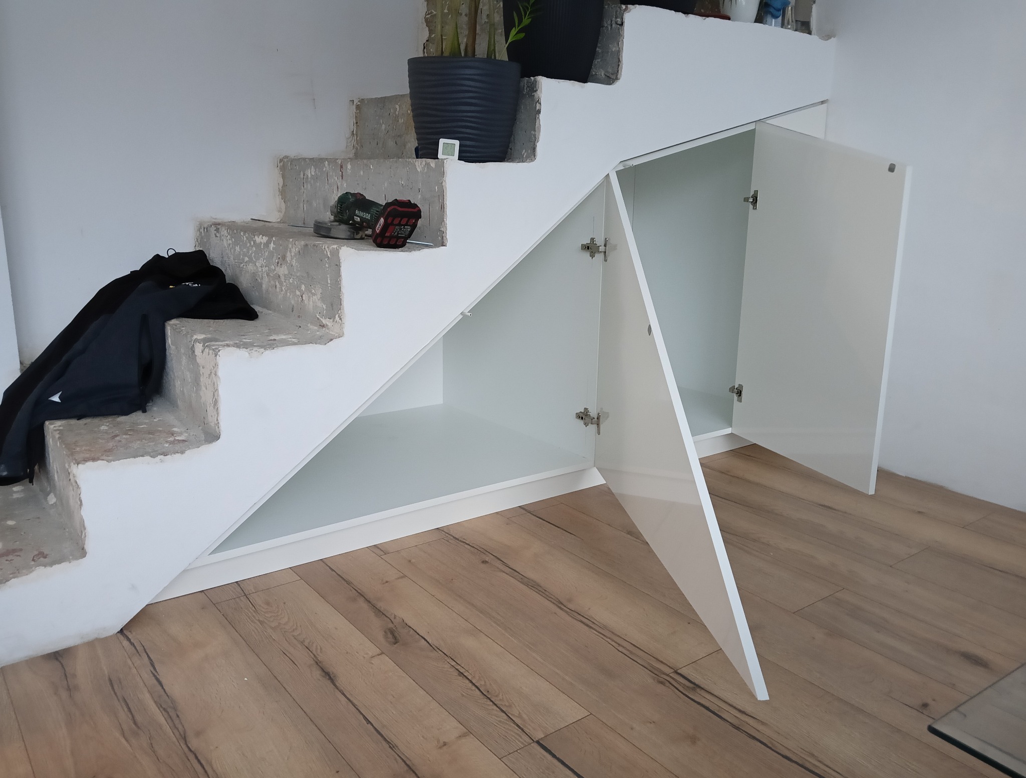 Biała szafka pod schodami z otwartymi drzwiczkami, na schodach narzędzia i ubrania, drewniana podłoga. Funkcjonalne wykorzystanie przestrzeni pod schodami.