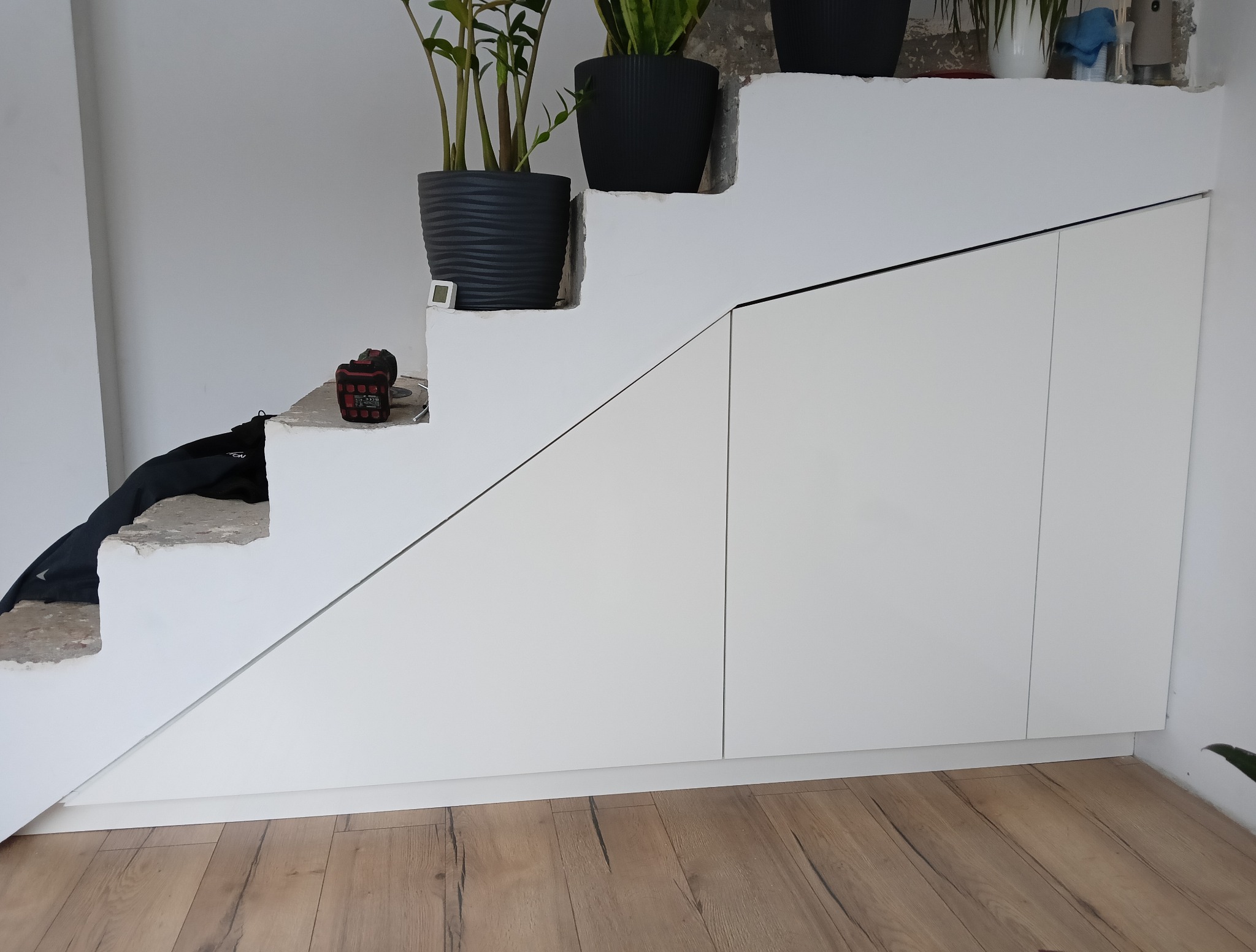 Białe szafki pod schodami z roślinami w doniczkach na stopniach, drewniana podłoga. Funkcjonalne wykorzystanie przestrzeni pod schodami.