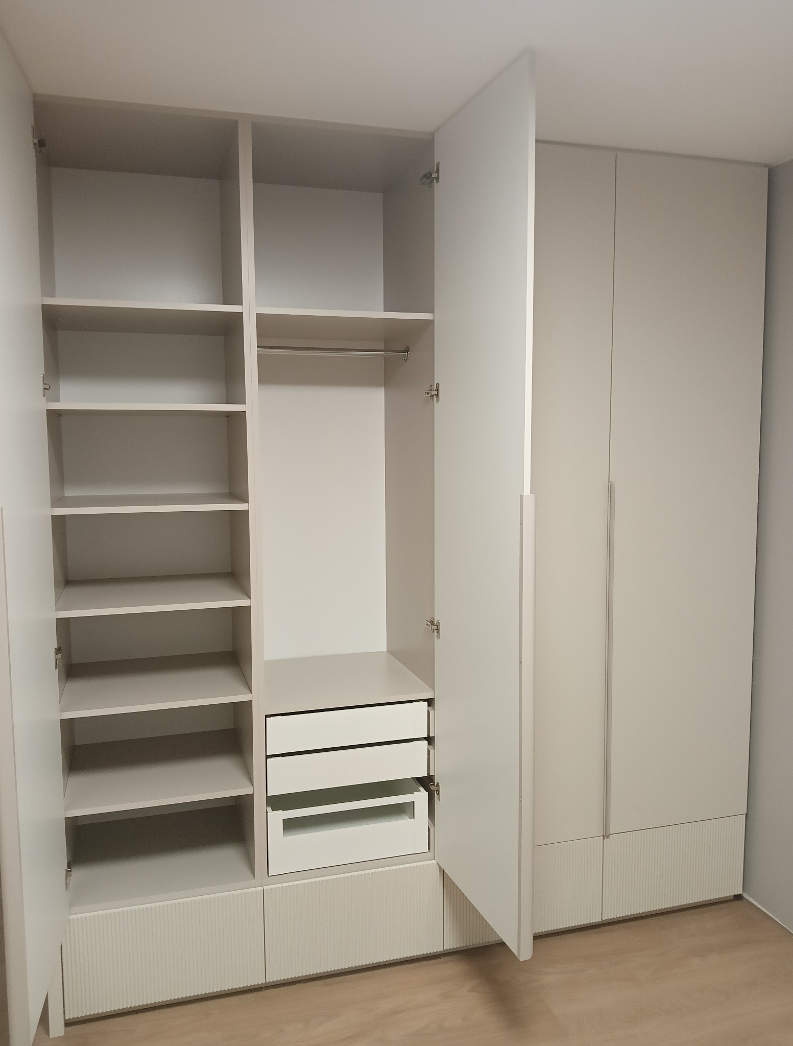 Jasna szafa wnękowa z otwartymi drzwiami, ukazująca półki, drążek na ubrania i szuflady. Dolna część szafy zdobiona pionowymi żłobieniami. Widoczna podłoga z paneli.
