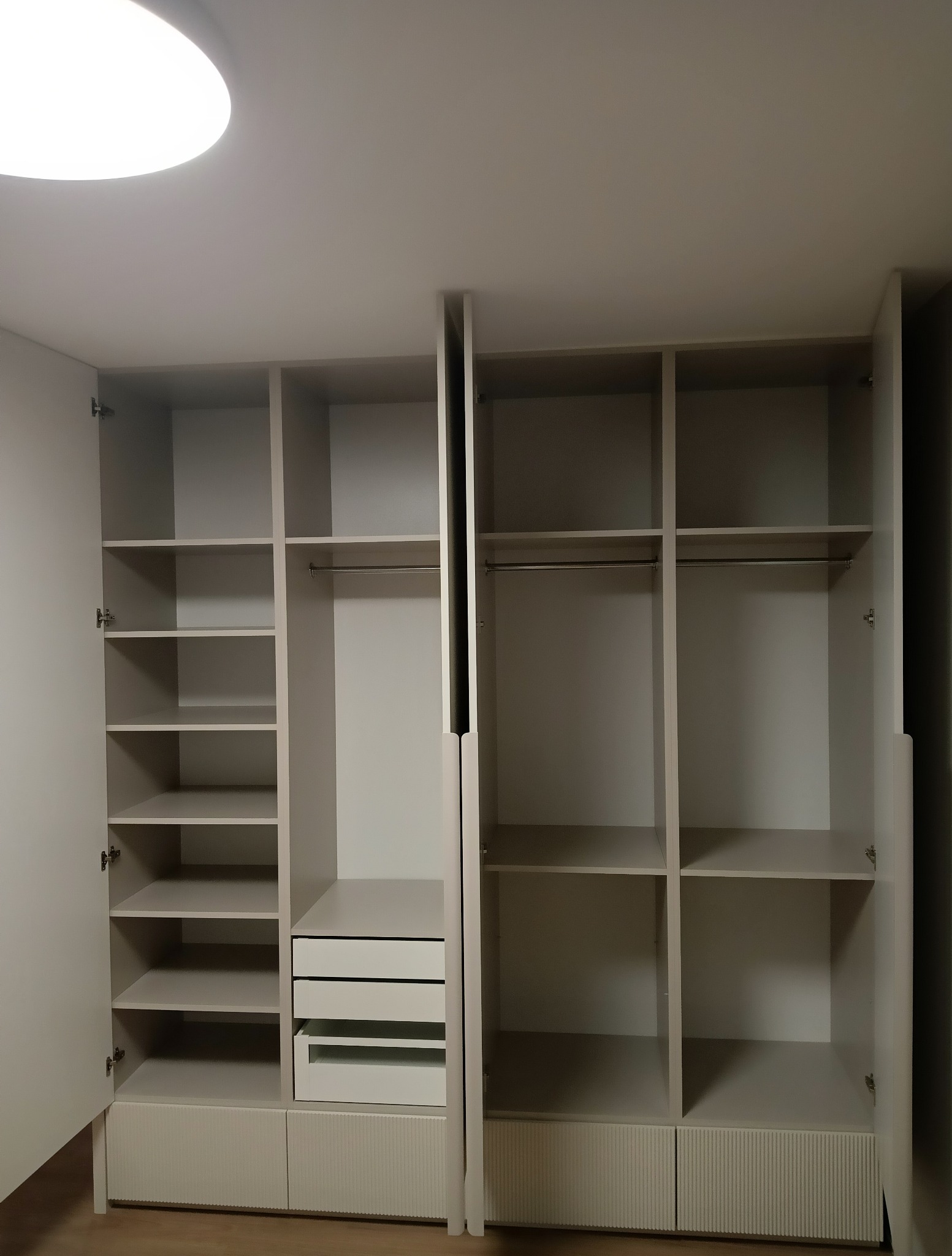 Jasnoszara szafa wnękowa z otwartymi drzwiami, widoczne półki, szuflady i drążki na ubrania. Minimalistyczny design, subtelne frezowanie na frontach dolnych szafek.