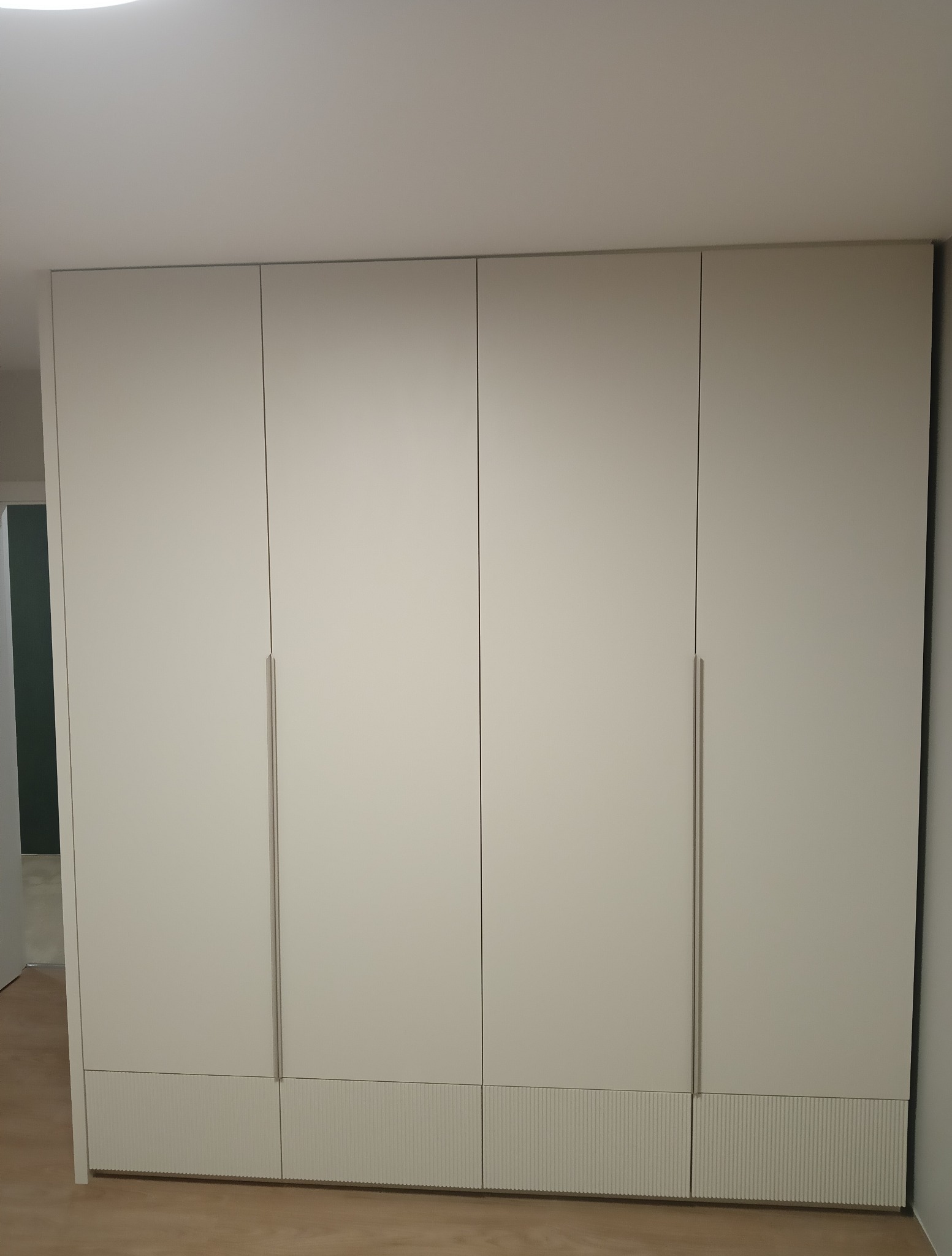 Czterodrzwiowa szafa wnękowa w odcieniach bieli z subtelnymi, pionowymi żłobieniami u dołu, minimalistyczne uchwyty, widok z przodu na tle jasnego wnętrza.