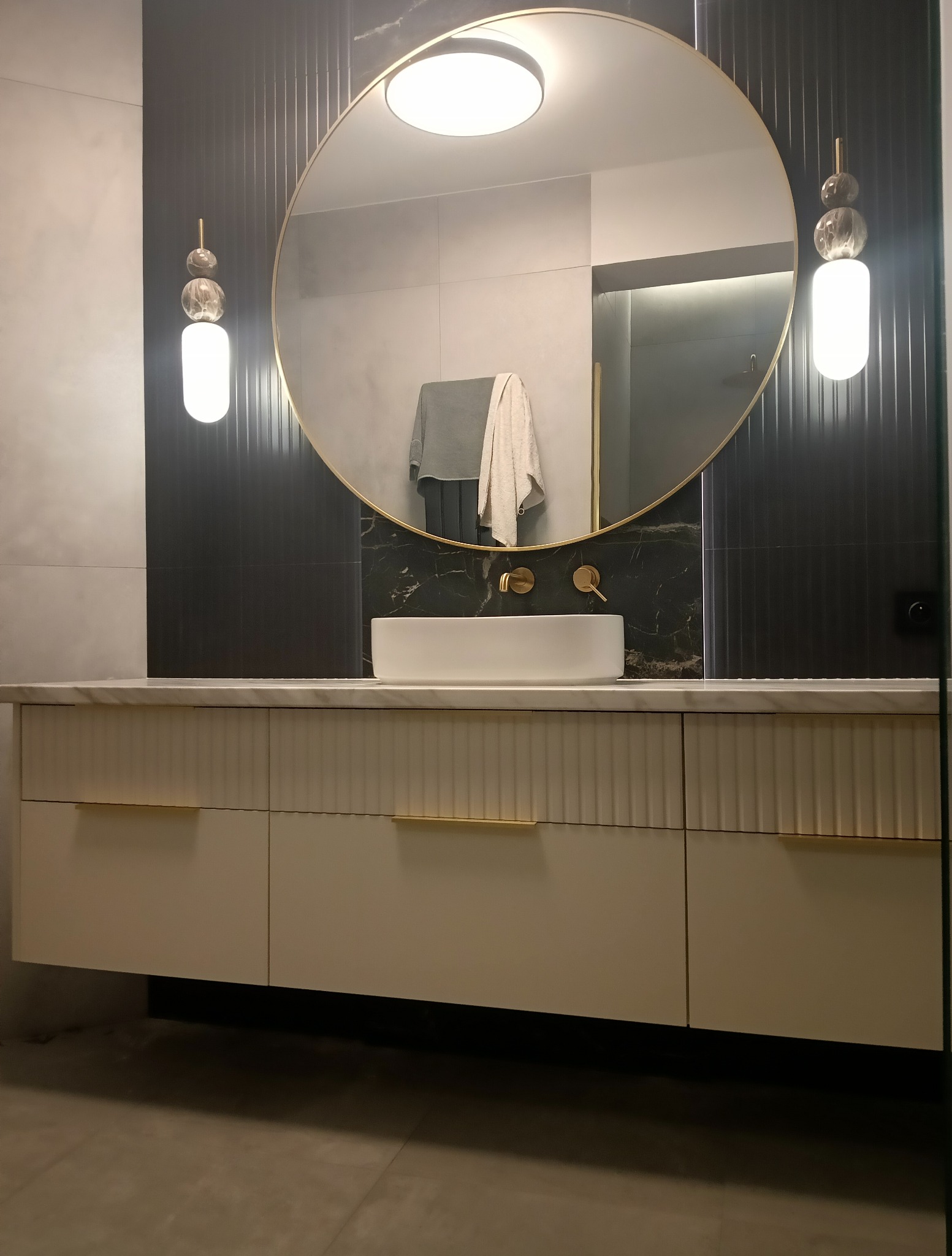 Nowoczesna łazienka z owalnym lustrem w złotej ramie, białą umywalką nablatową i szafką z frezowanymi frontami oraz złotymi uchwytami. Minimalistyczne oświetlenie.