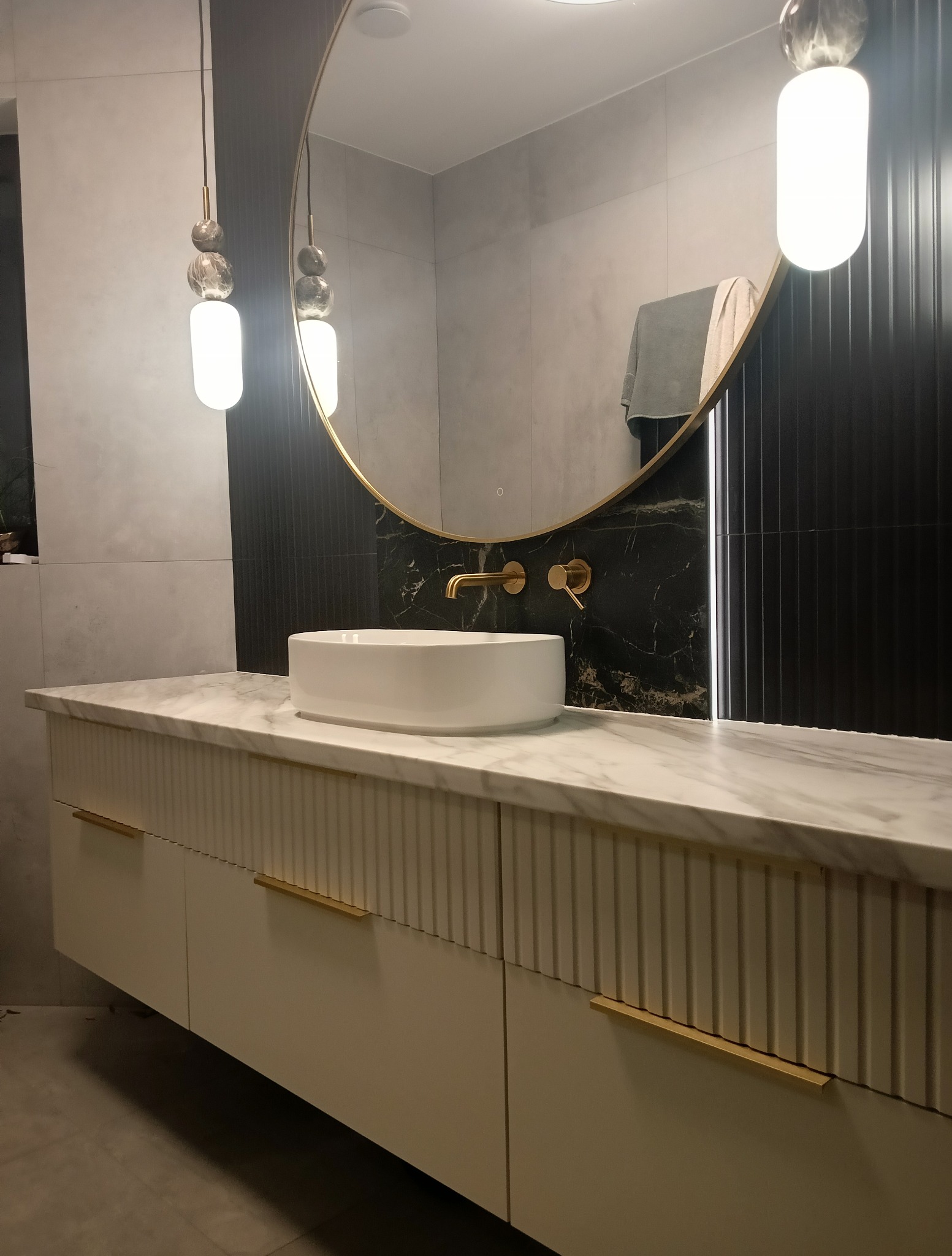 Nowoczesna łazienka z owalnym lustrem w złotej ramie, białą umywalką nablatową i szafką z frezowanymi frontami oraz marmurowym blatem. Złota armatura i designerskie lampy.