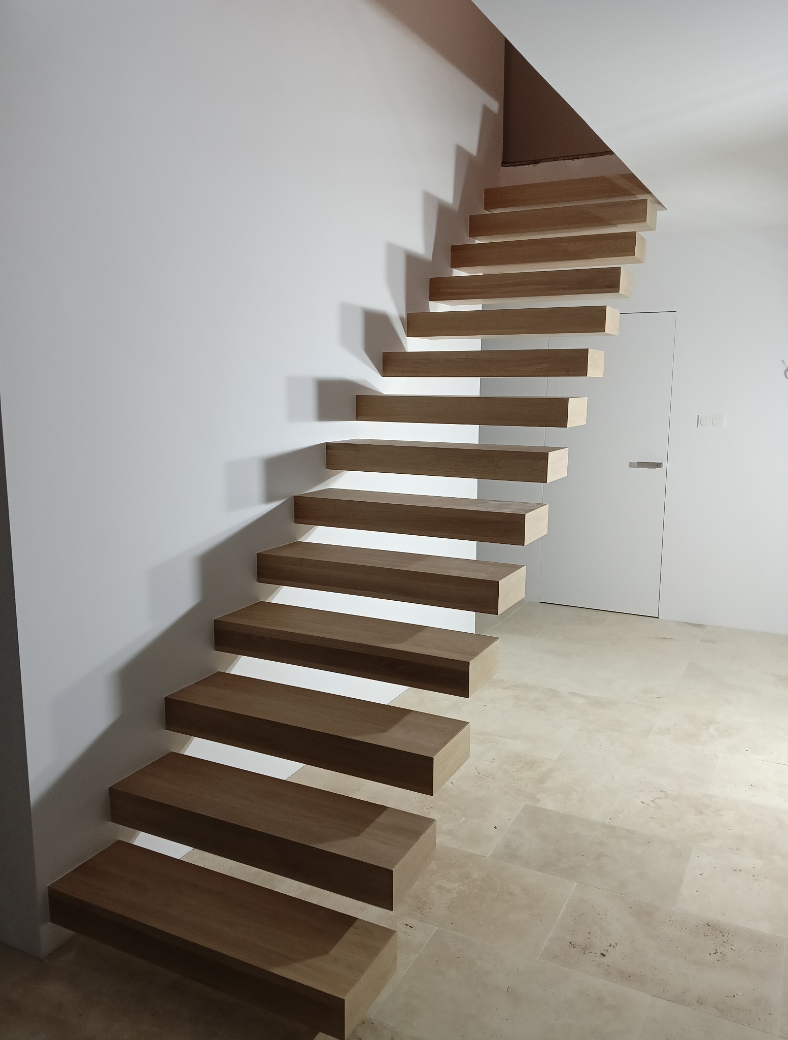Nowoczesne, drewniane schody wspornikowe zamontowane przy białej ścianie, z widoczną fakturą kamiennej podłogi i minimalistycznymi drzwiami w tle.