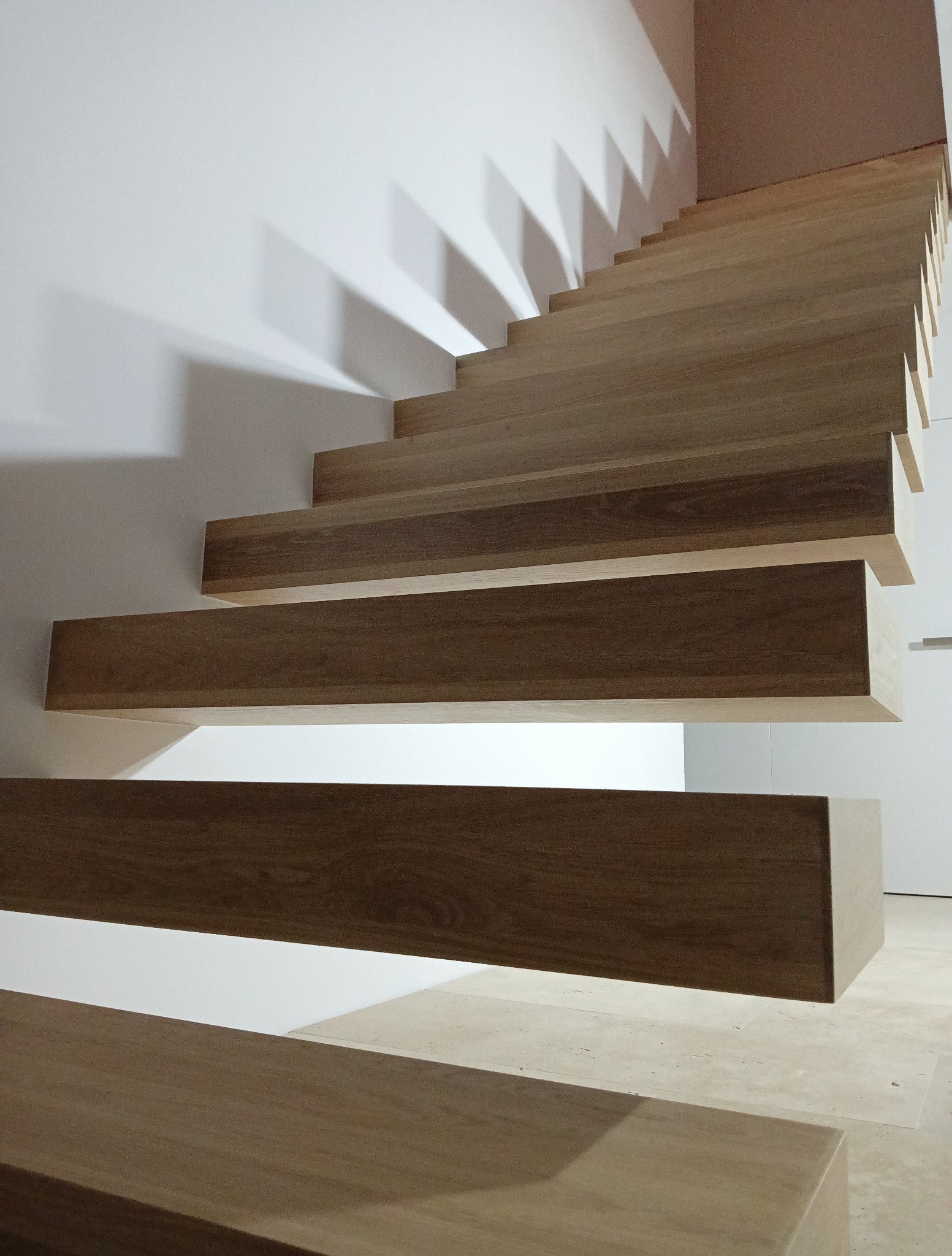 Nowoczesne, drewniane schody wspornikowe z subtelnym oświetleniem, rzucające geometryczne cienie na białą ścianę. Minimalistyczny design i ciepła kolorystyka drewna.