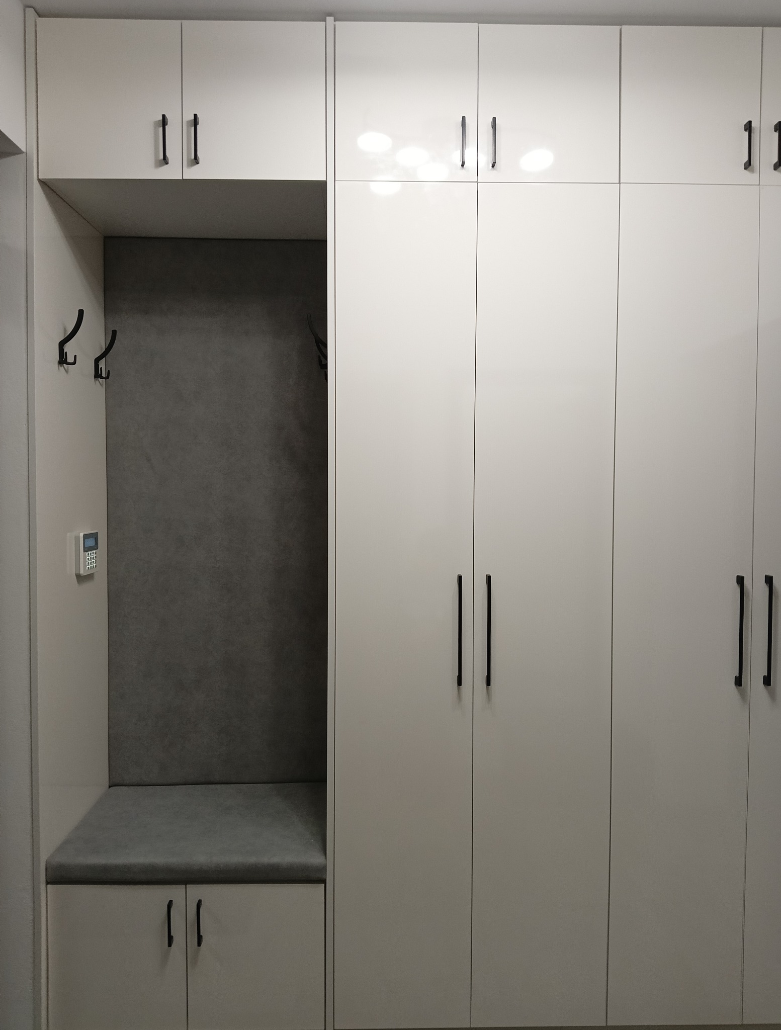 Biała szafa wnękowa z siedziskiem i wieszakami, czarne uchwyty, szara tapicerka siedziska i panelu ściennego. Nowoczesny design, minimalistyczny styl.