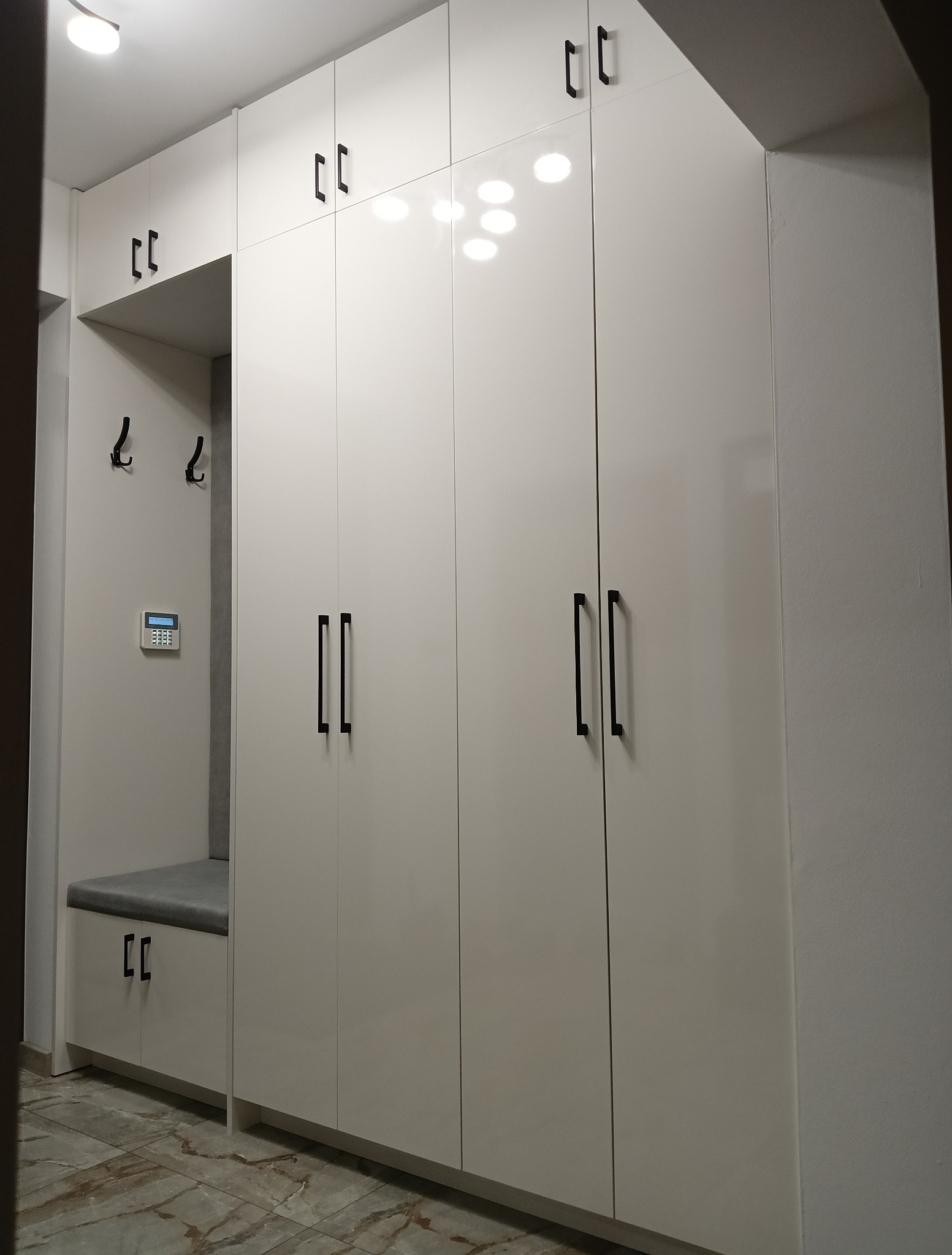 Biała szafa wnękowa z siedziskiem i wieszakami w przedpokoju. Minimalistyczne czarne uchwyty, marmurowa podłoga. Widoczny panel alarmowy.