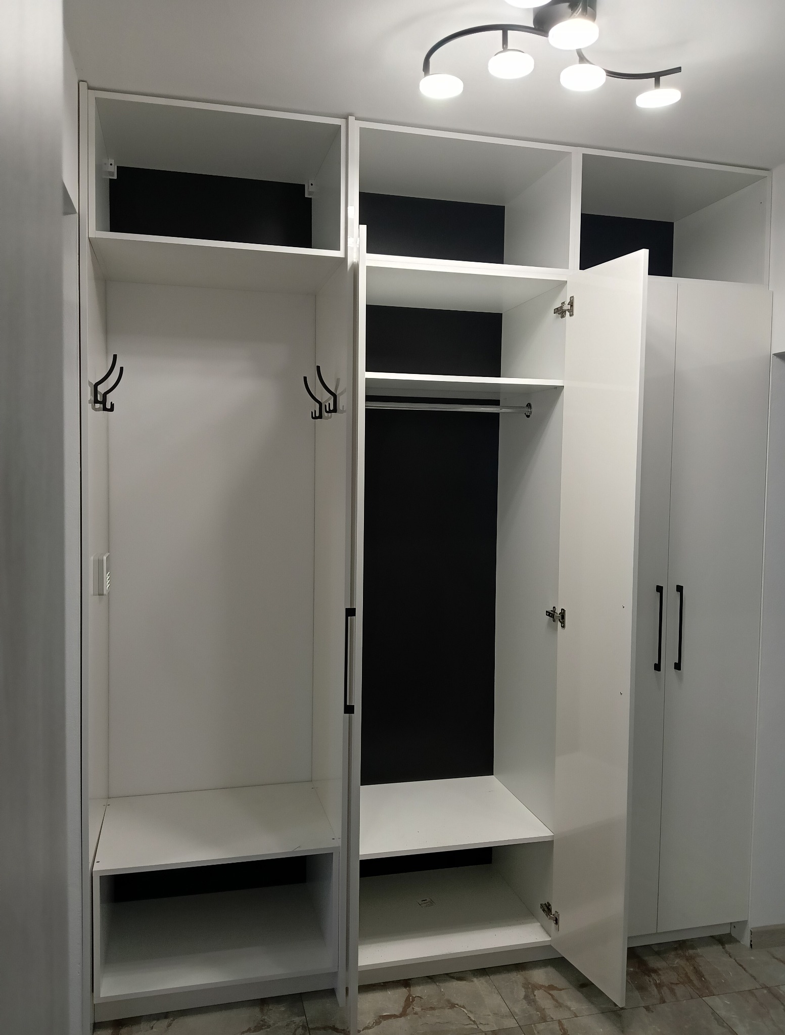 Biała szafa wnękowa z czarnym tłem, otwarte drzwi ukazują półki i wieszaki. Minimalistyczny design, nowoczesne oświetlenie sufitowe. Widoczne zawiasy i uchwyty.