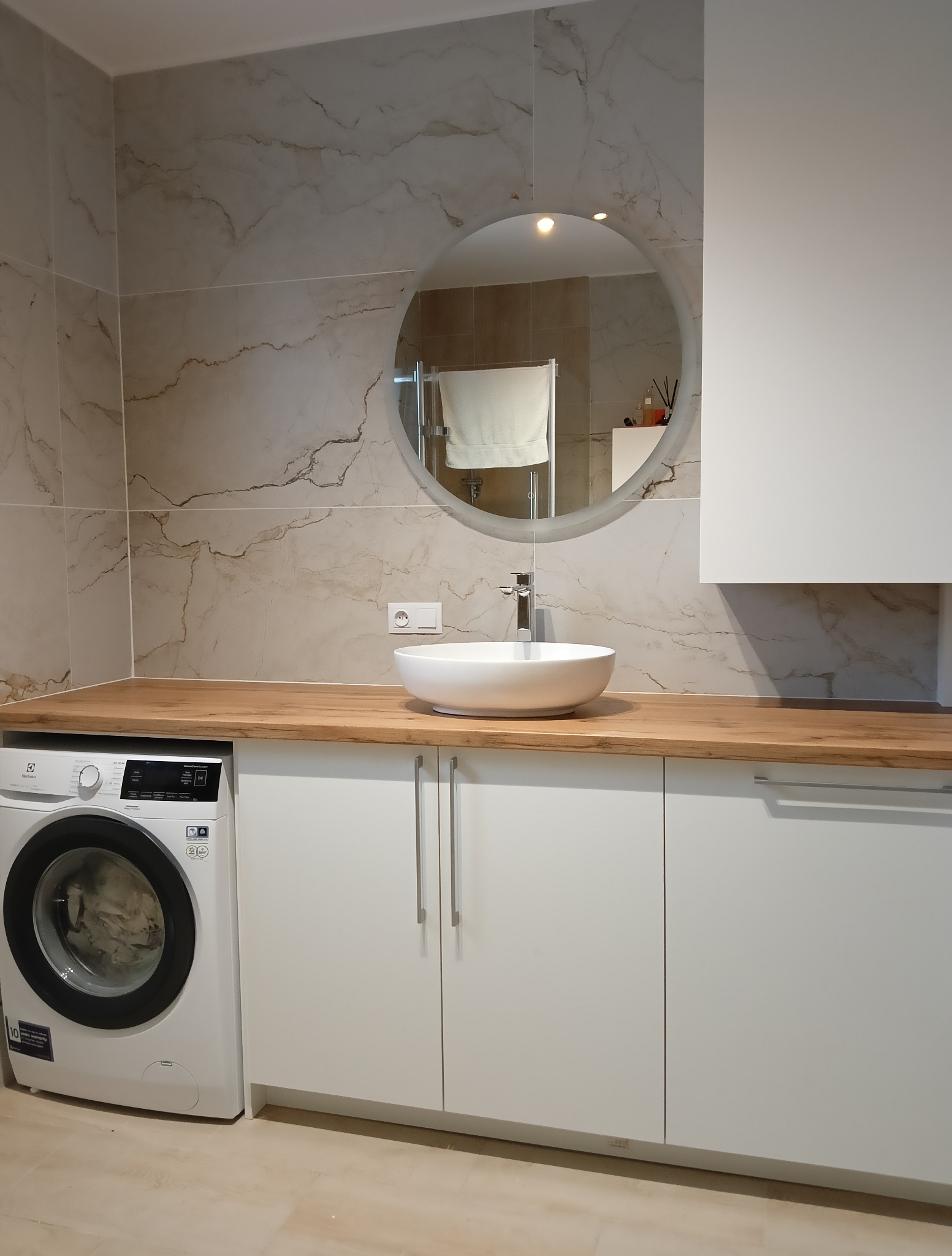 Nowoczesna łazienka z drewnianym blatem, okrągłym lustrem i białą umywalką nablatową. Pralka Electrolux wkomponowana w zabudowę. Marmurowe płytki na ścianie.