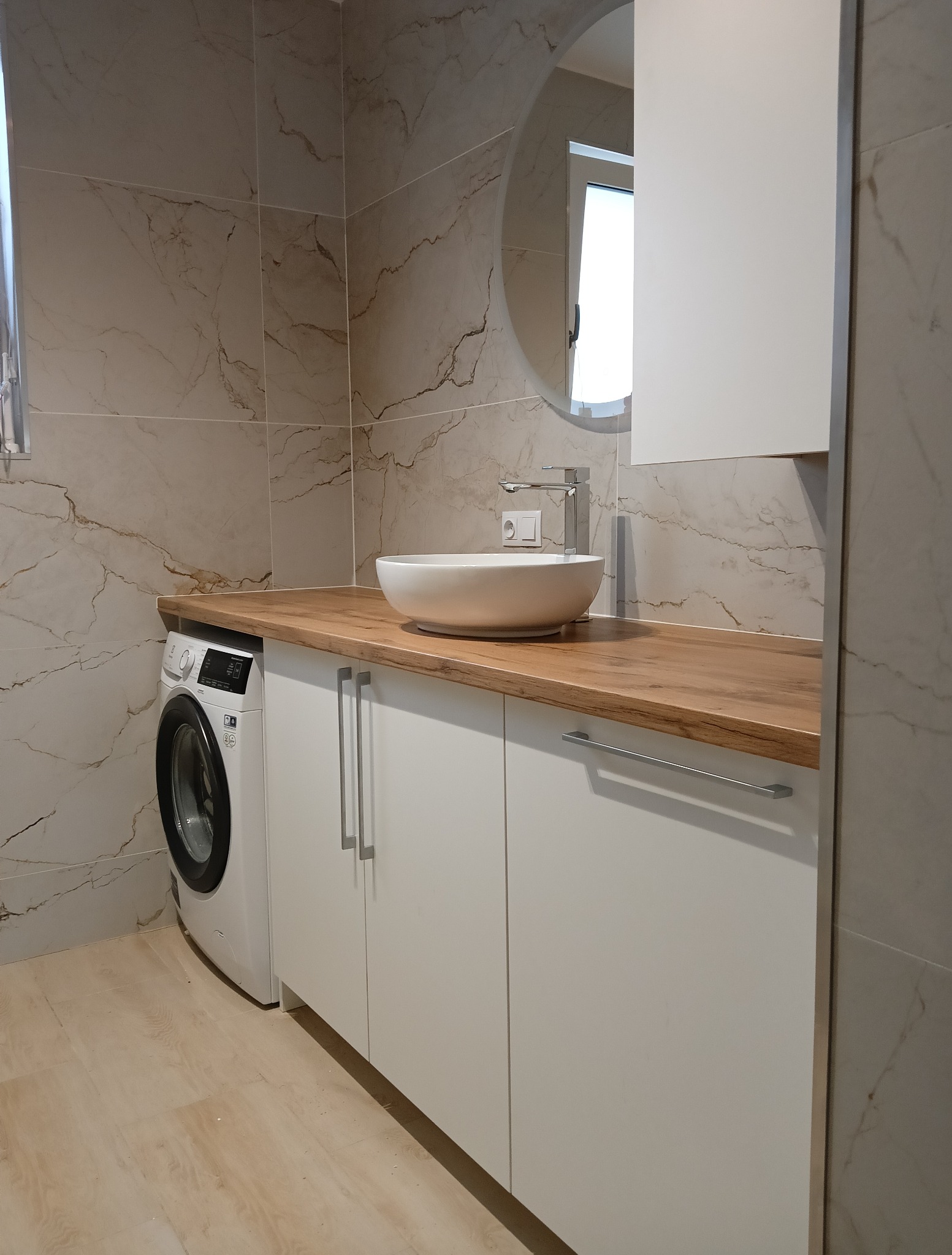 Nowoczesna łazienka z drewnianym blatem, białą umywalką nablatową i szafkami. Obok pralka, a na ścianie lustro w okrągłej ramie i marmurowe płytki.