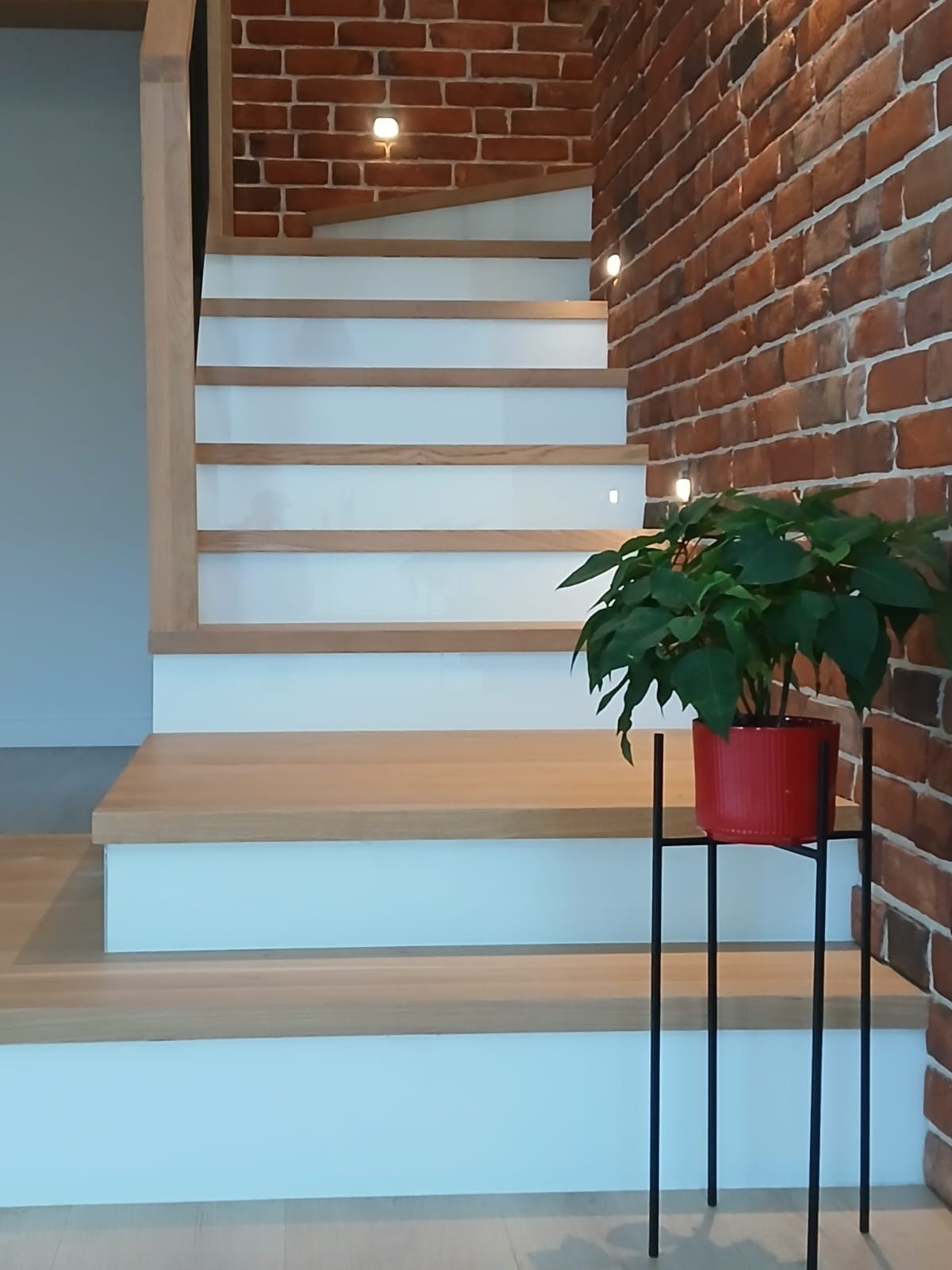 Nowoczesne schody drewniane z białymi podstopnicami i oświetleniem punktowym na ceglanej ścianie. Obok czerwona doniczka z zieloną rośliną na metalowym stojaku.