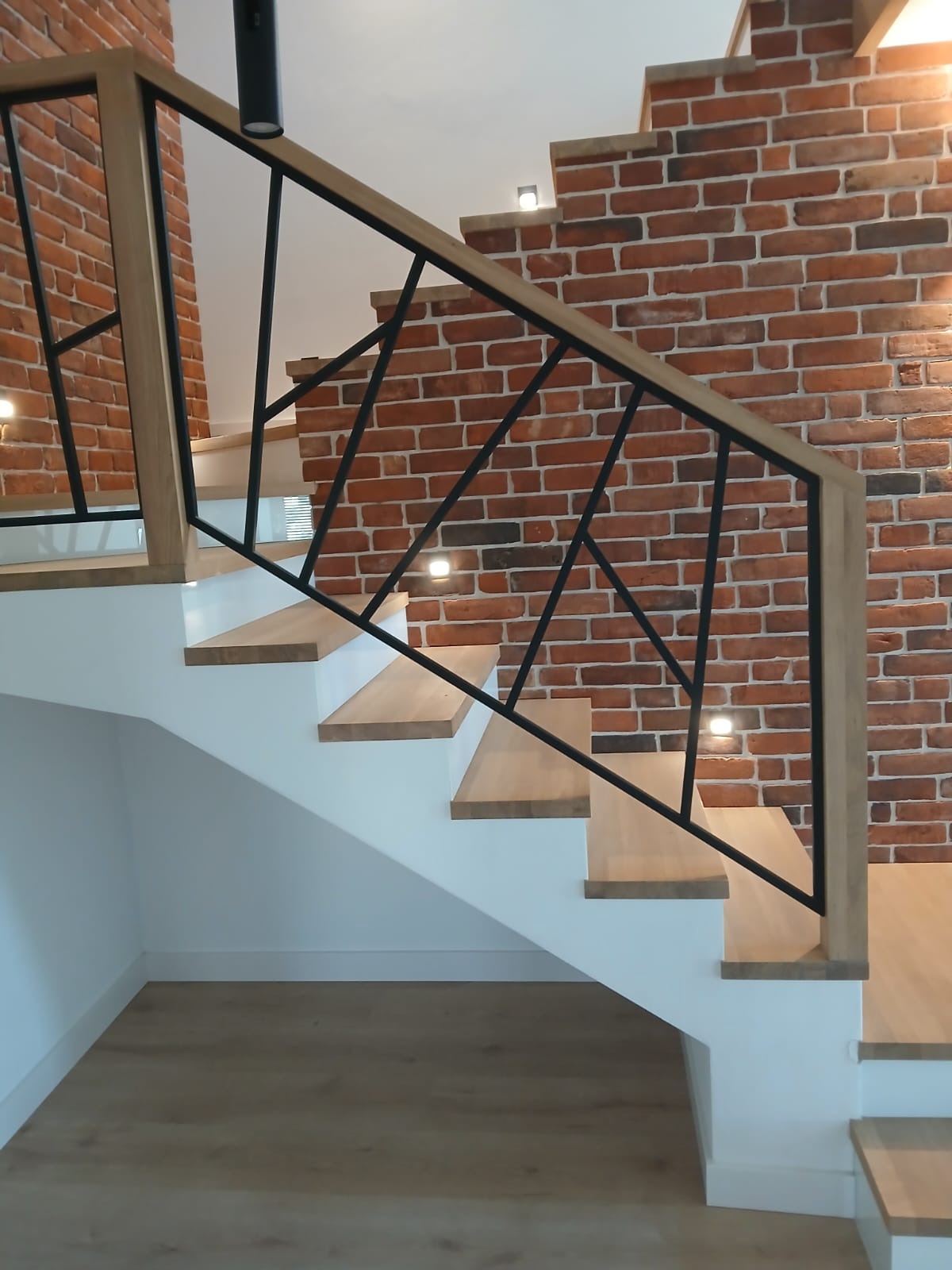 Nowoczesne schody drewniane z czarną, geometryczną balustradą na tle ściany z czerwonej cegły, oświetlone punktowymi lampkami. Jasne stopnie i białe podstopnice.