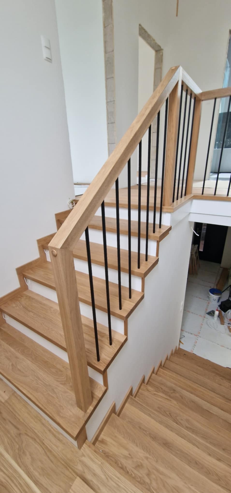 Jasne, drewniane schody z czarnymi, metalowymi balustradami w nowoczesnym wnętrzu. Widoczne niedokończone wykończenia ścian.